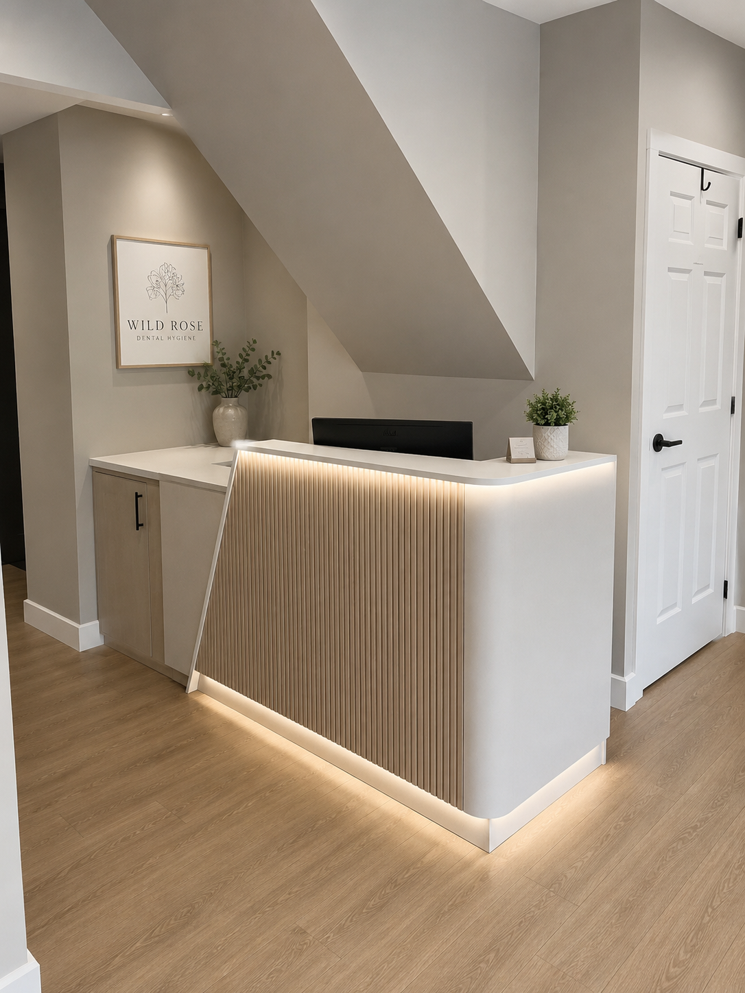 Modern dental reception area with a white and wooden countertop, decorative plants, and a sign that reads 'Wild Rose Dental Hygiene' on the wall.