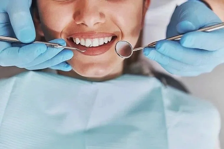 A person in a dental chair smiling with a dental mirror and tweezers, wearing blue gloves, during a dental examination or procedure.
