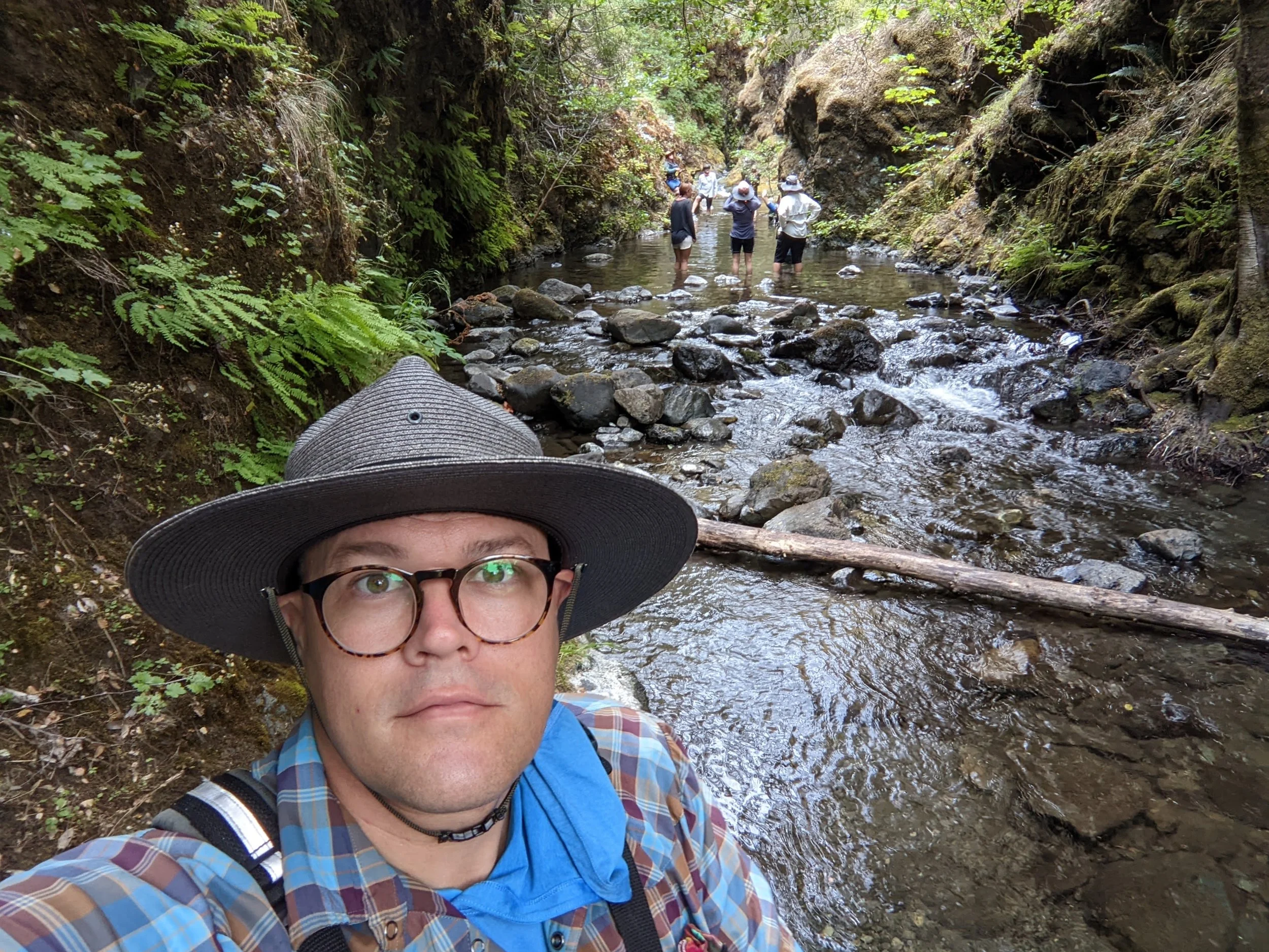Person taking a selfie in a lush green forest stream with other people in the water in the background.