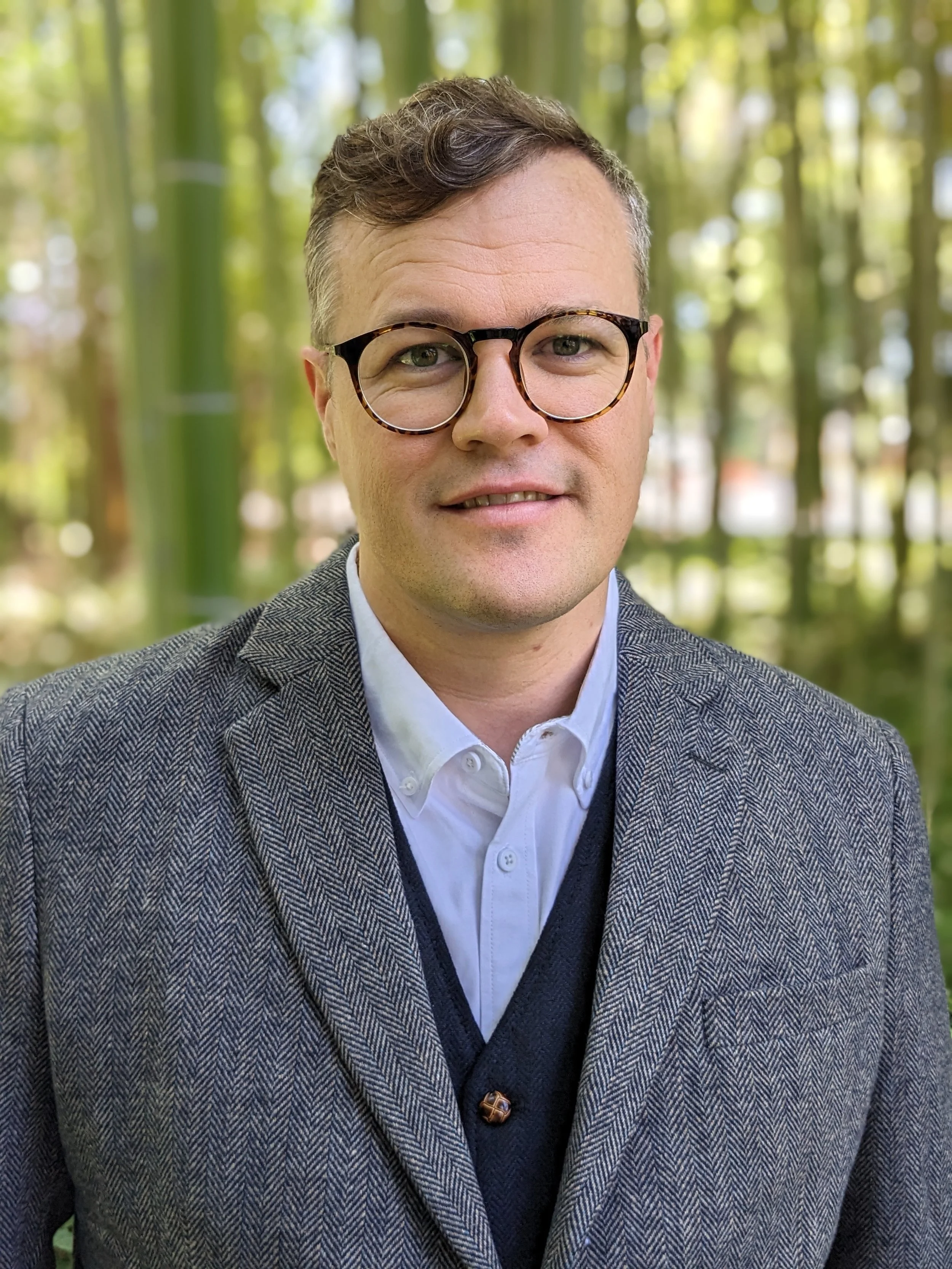 A close-up portrait of a man with glasses, wearing a white shirt, dark vest, and patterned blazer, standing outdoors in a green, wooded area.