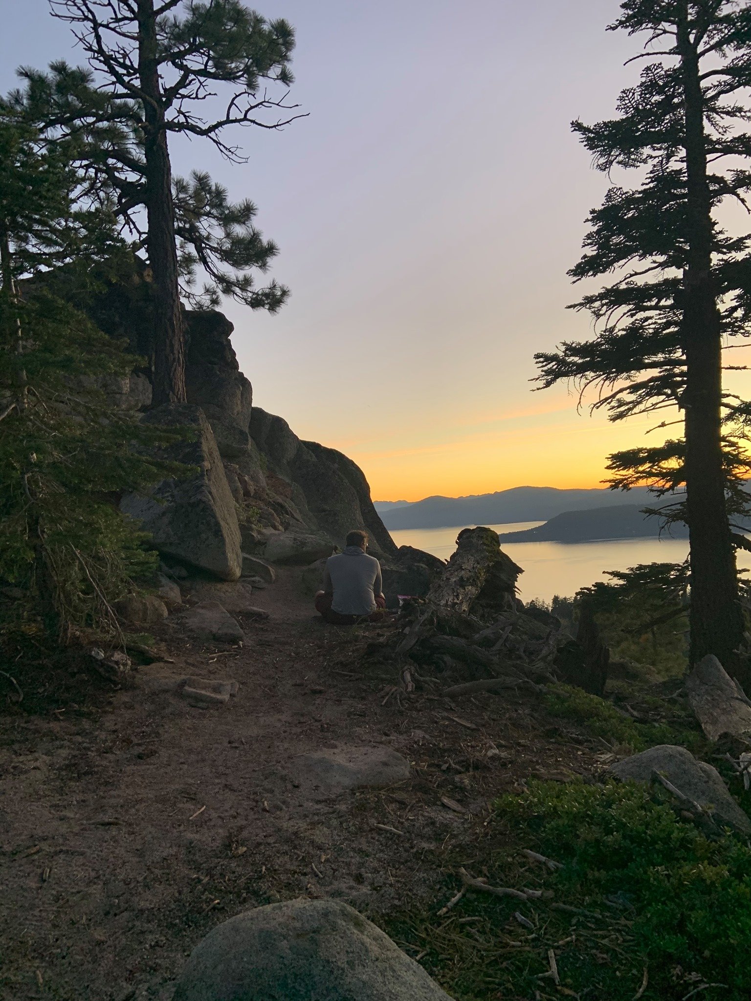 A person sitting on the ground near large rocks and trees, overlooking a body of water with mountains in the distance at sunset.