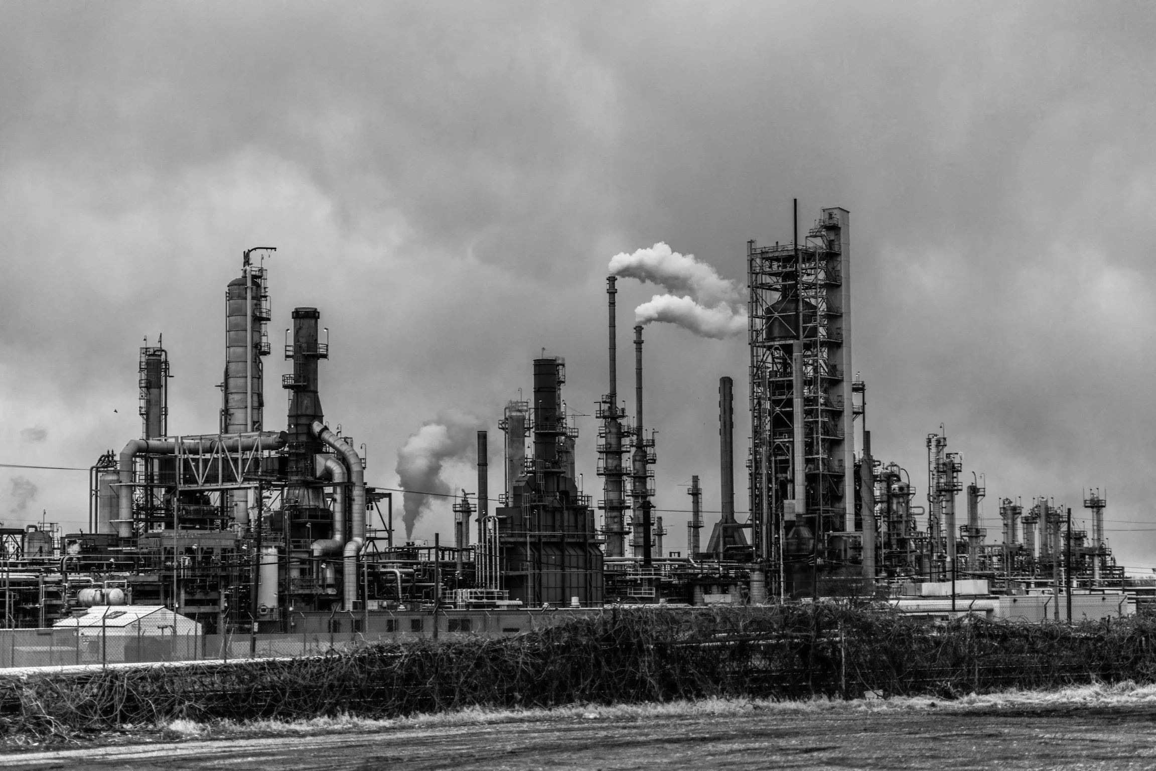 Industrial oil refinery with tall processing towers emitting white smoke under cloudy sky.