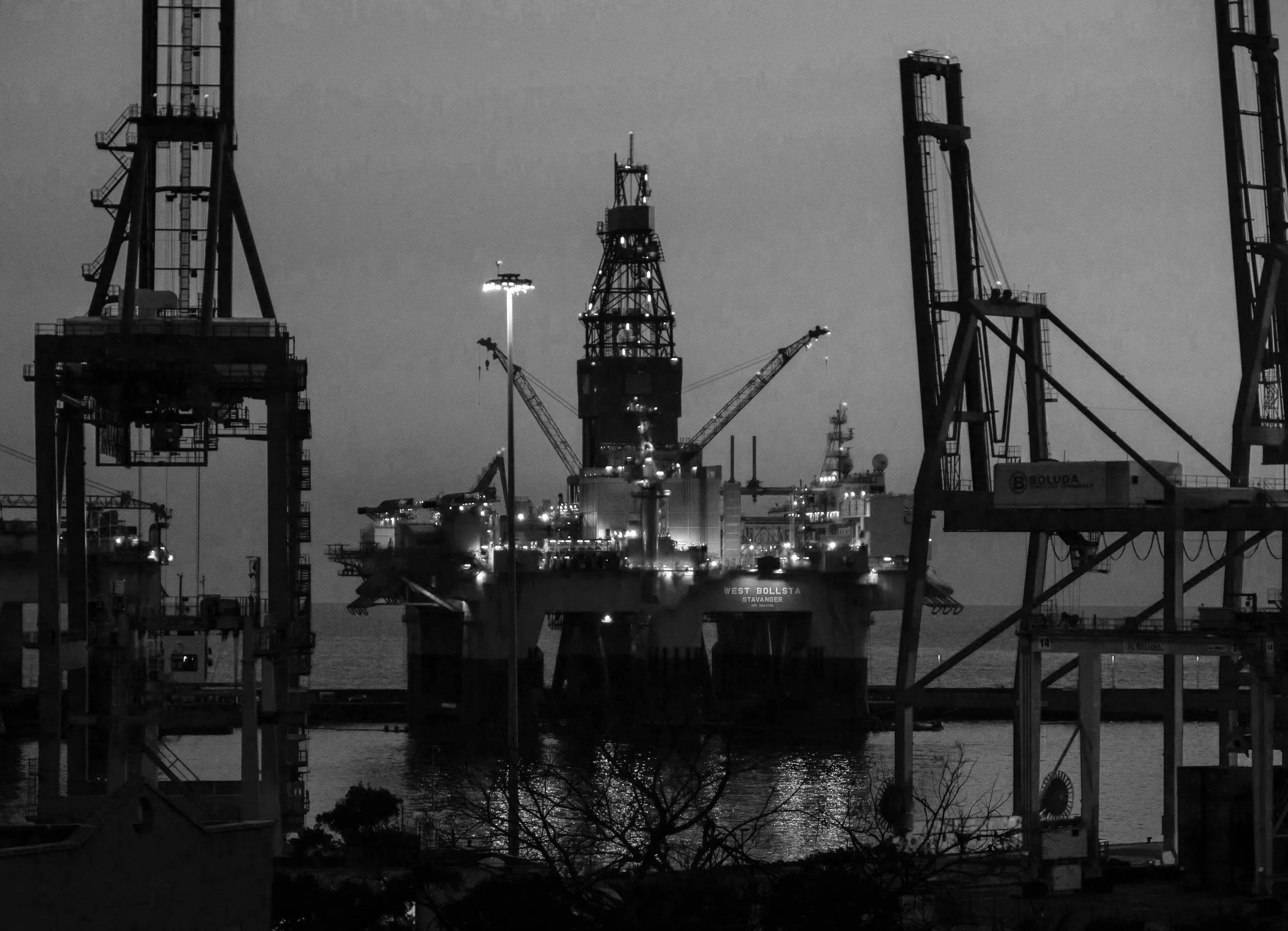 Nighttime view of an offshore oil drilling platform with multiple cranes and tall structures, illuminated by artificial lights, near a body of water with trees in the foreground.