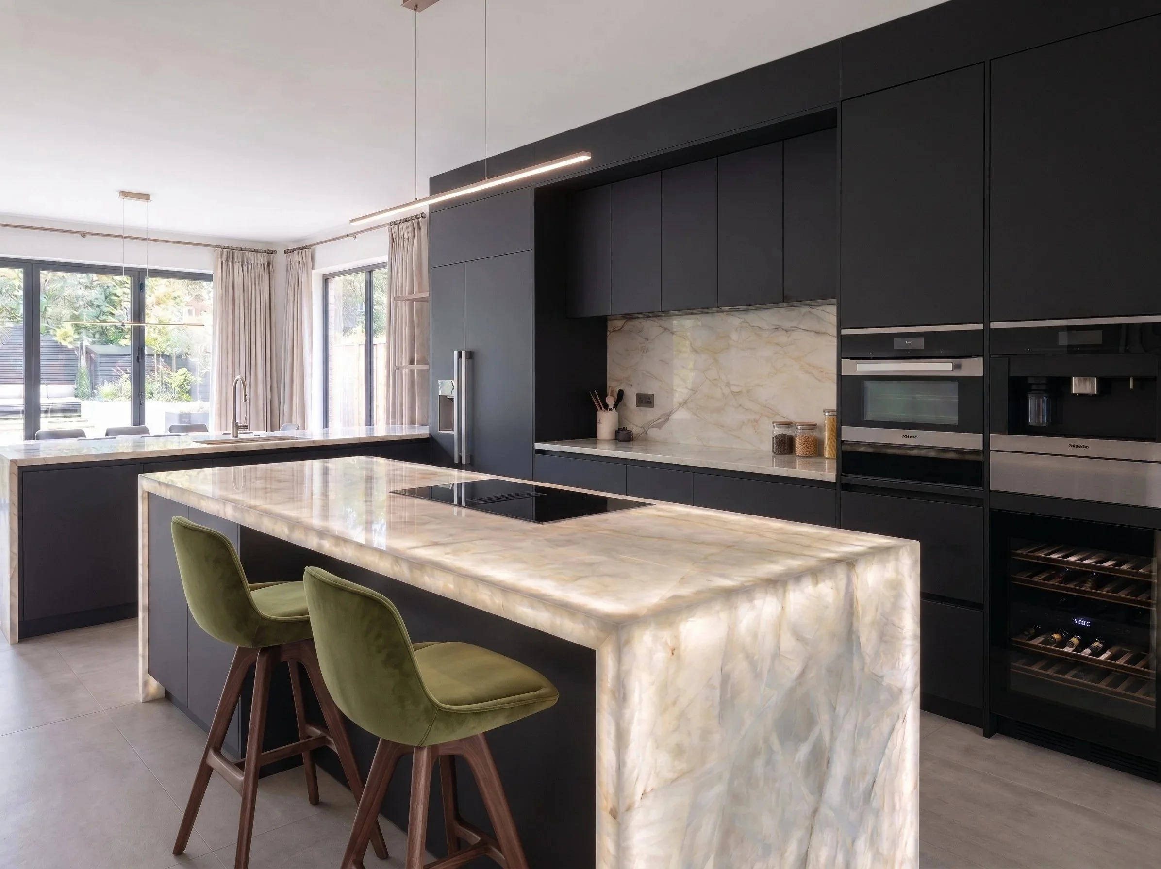 Cocina moderna de diseño minimalista con armarios oscuros, encimeras de mármol claro, ventanas grandes con cortinas beige y una isla central con sillas verdes.