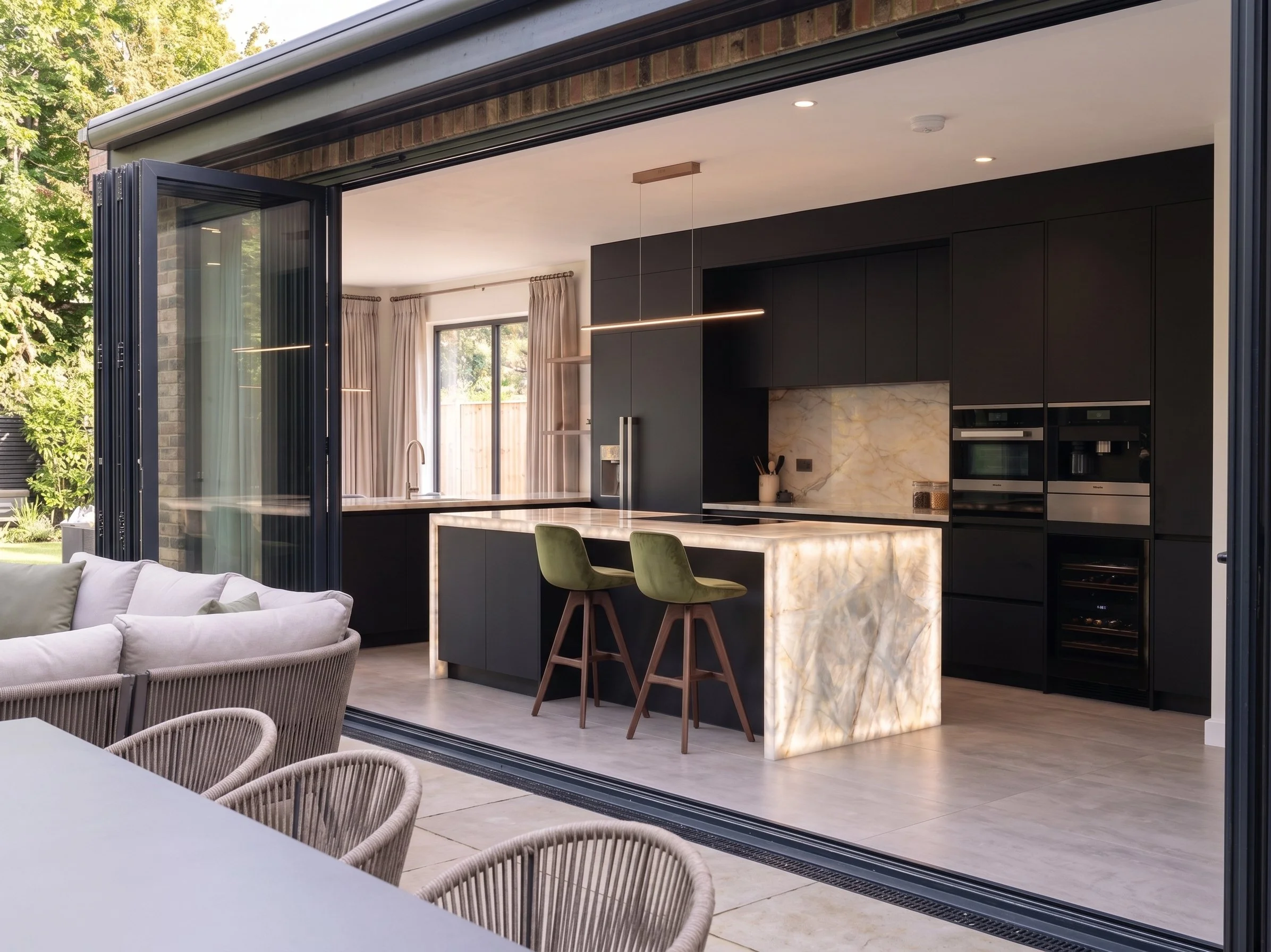 interior de una cocina moderna vista desde un espacio exterior con puertas plegables abiertas, con una isla de mármol y sillas verdes