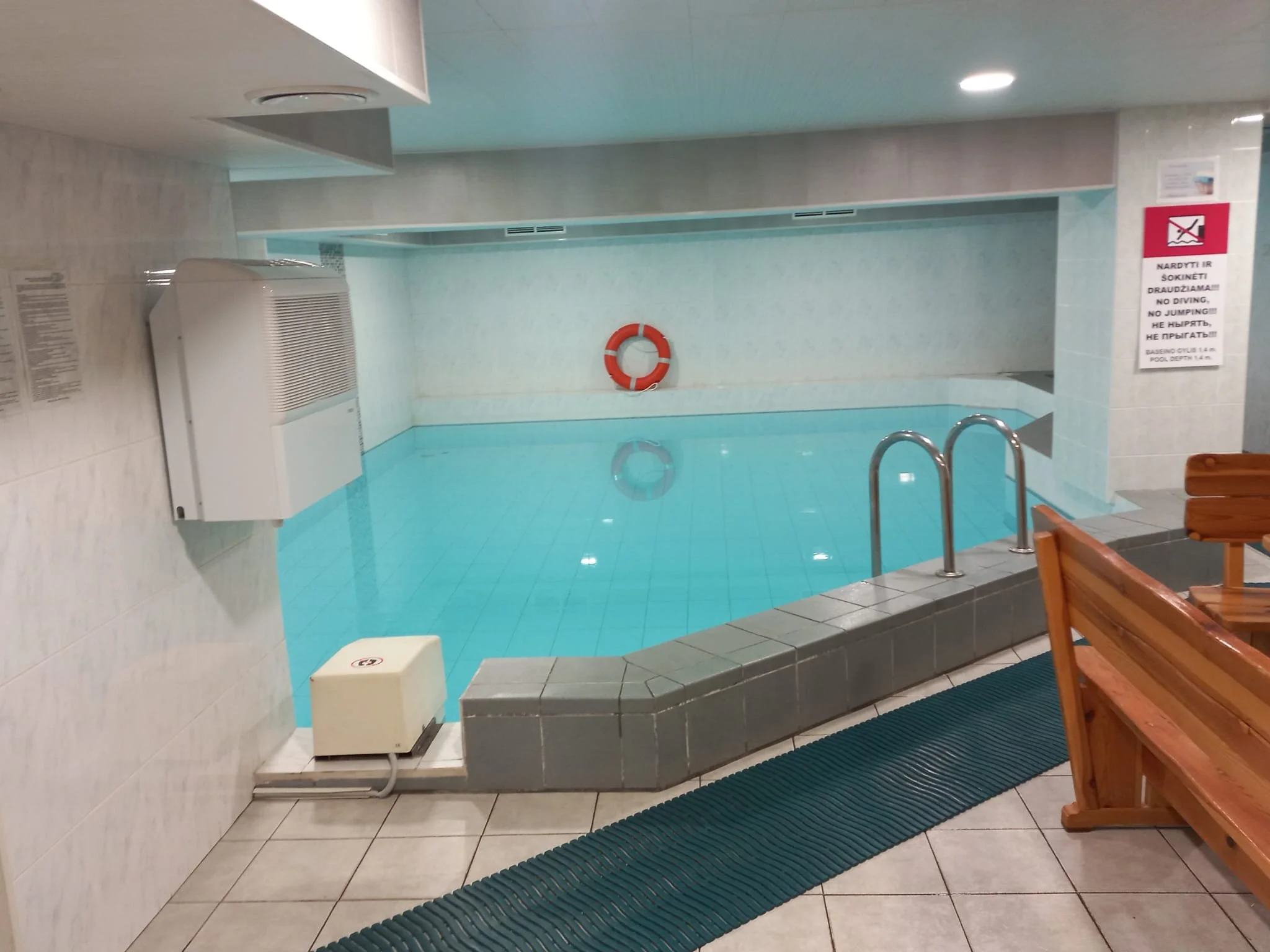 Bilder på ett inomhusbad med ett blått pool, räddningsring på väggen, trappa och träbänkar. En skylt med varningsmeddelande om drunkning.