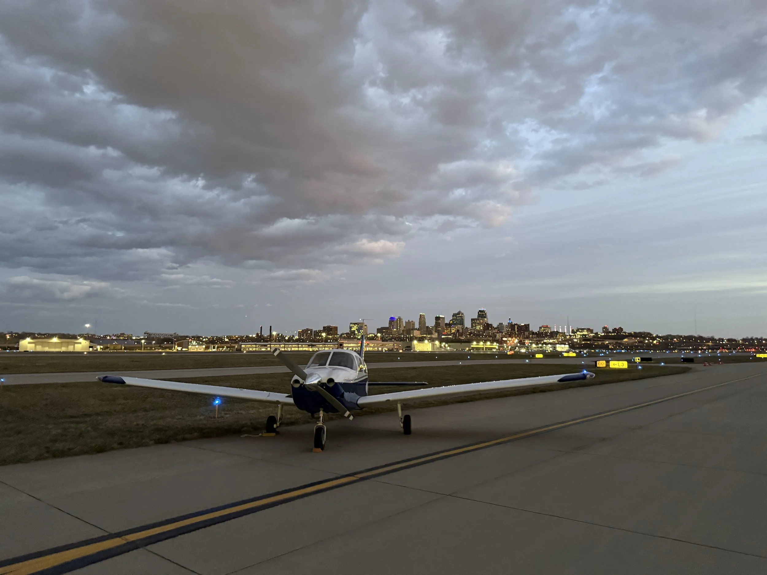 Aviation training airplane on tarmac with a city skyline in the background, under a cloudy sky during dusk.