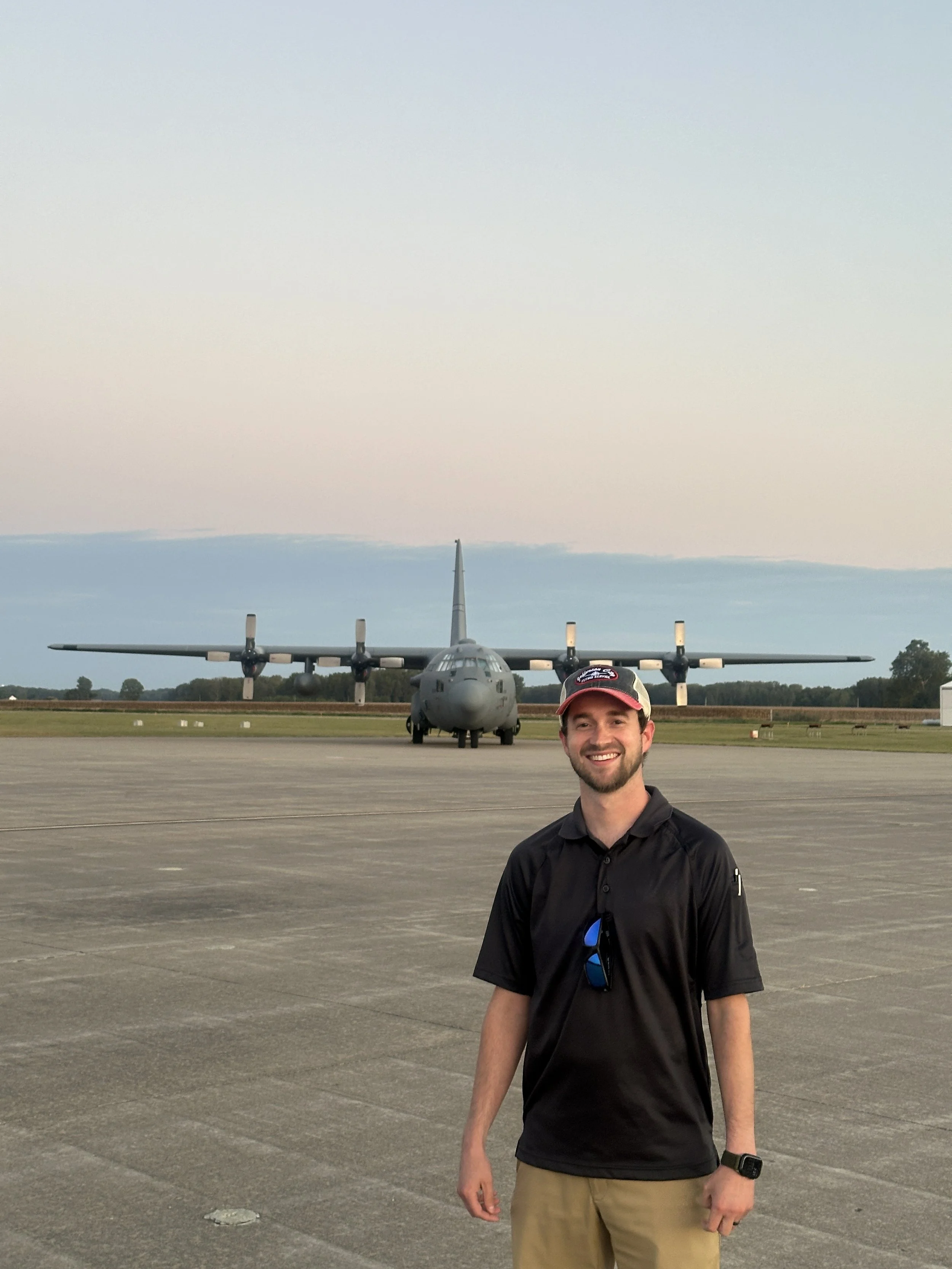 A smiling certified flight instructor in Springfield Missouri area with a beard, wearing a black shirt, beige pants, a cap, and a watch, stands in front of a military cargo plane on an open tarmac at an airport during daytime.