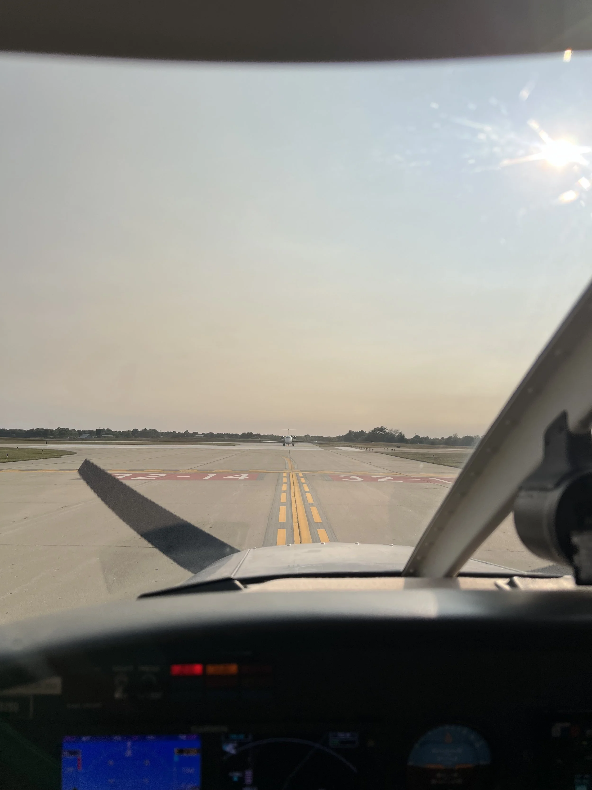 Student pilot view from inside an aircraft cockpit showing the runway ahead with a small airplane positioned on the runway, under a clear sky with sunlight.