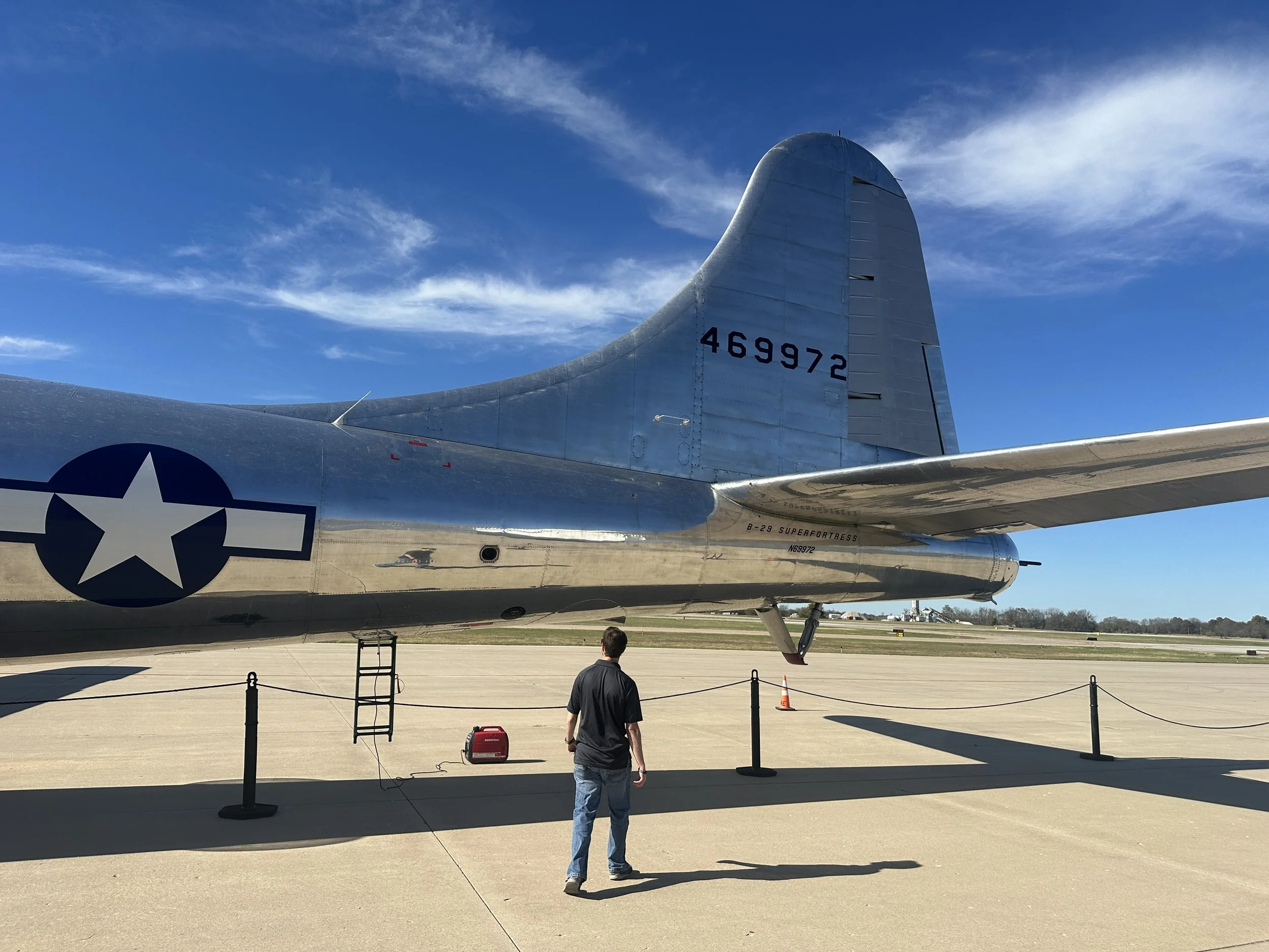 A vintage silver military aircraft with the markings of the United States Air Force, displayed outside on a sunny day with a clear blue sky, surrounded by a black rope barrier, with a person walking nearby.