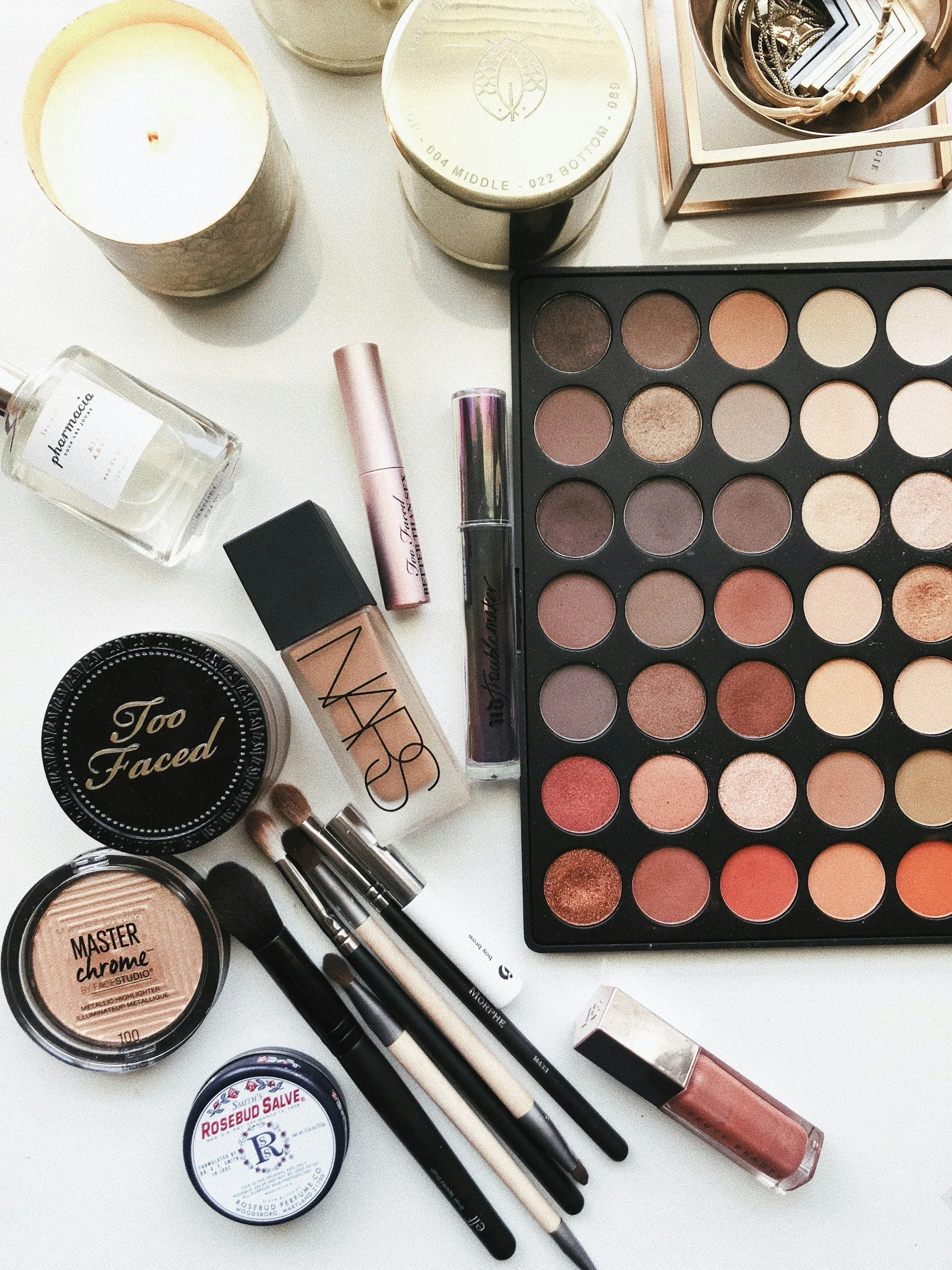 A flat lay of makeup products including eyeshadow palettes, brushes, foundation, lipstick, highlighter, setting spray, and candles.