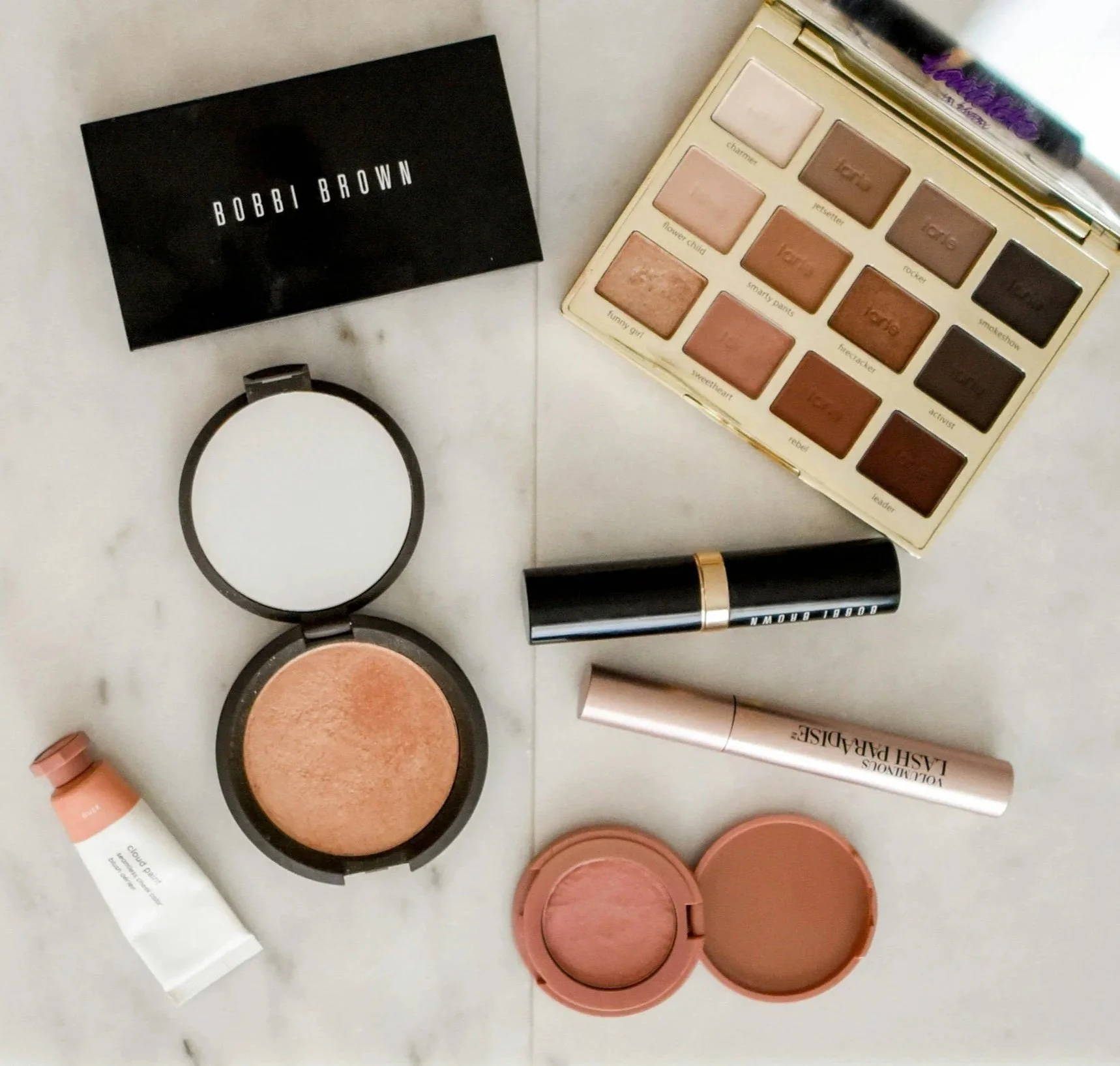 A collection of makeup products including eyeshadow palettes, blushes, highlighters, and lipsticks arranged on a white surface.