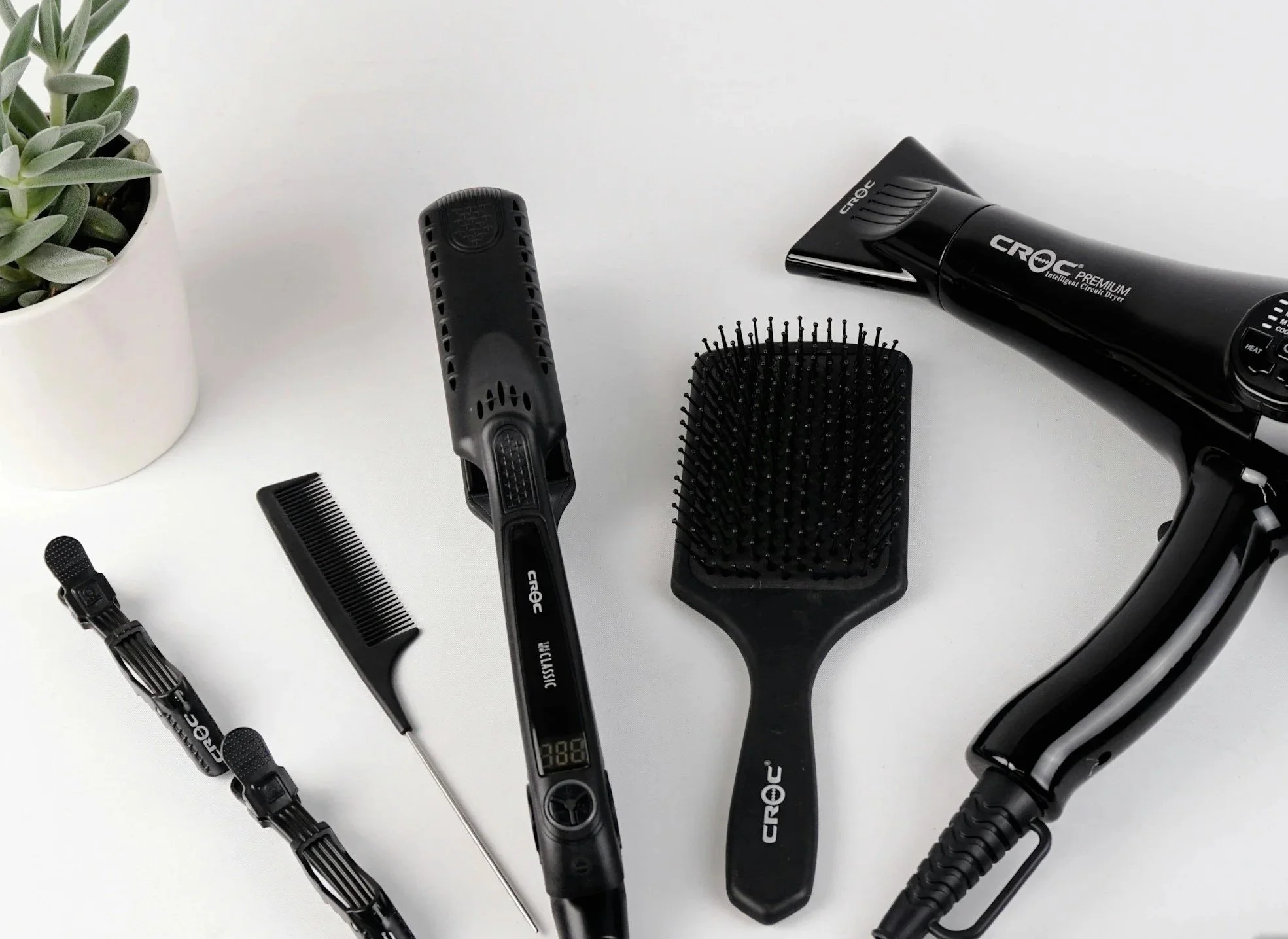 A white table with a potted succulent plant on the left and a collection of black hair styling tools on the right, including a hair dryer, a brush, a wide-tooth comb, and various hair clips.