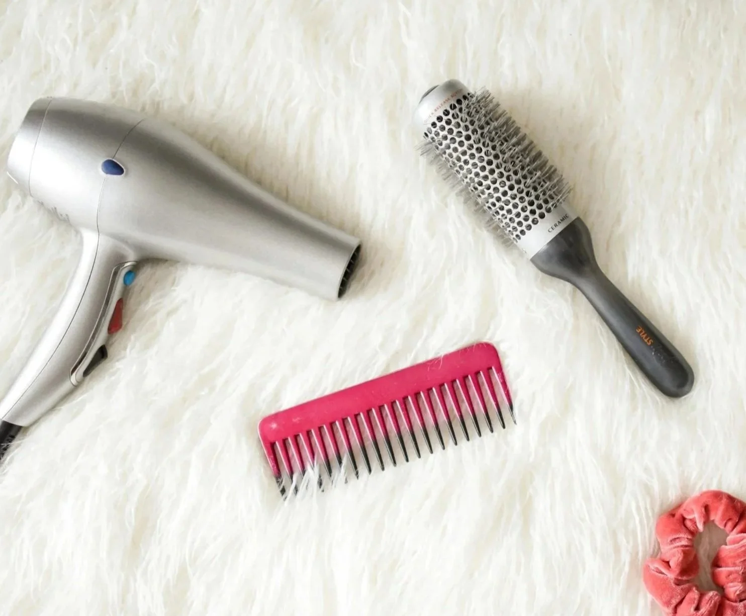 A hairdryer, a round hairbrush, and a pink hairbrush on a white fluffy surface with a pink scrunchie at the bottom right corner.