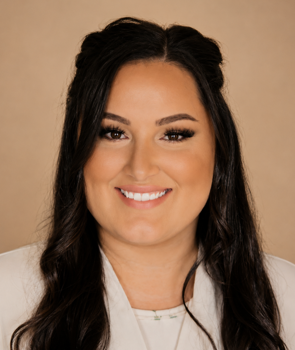 Close-up portrait of a woman with long dark wavy hair, smiling, wearing a white blazer and makeup.
