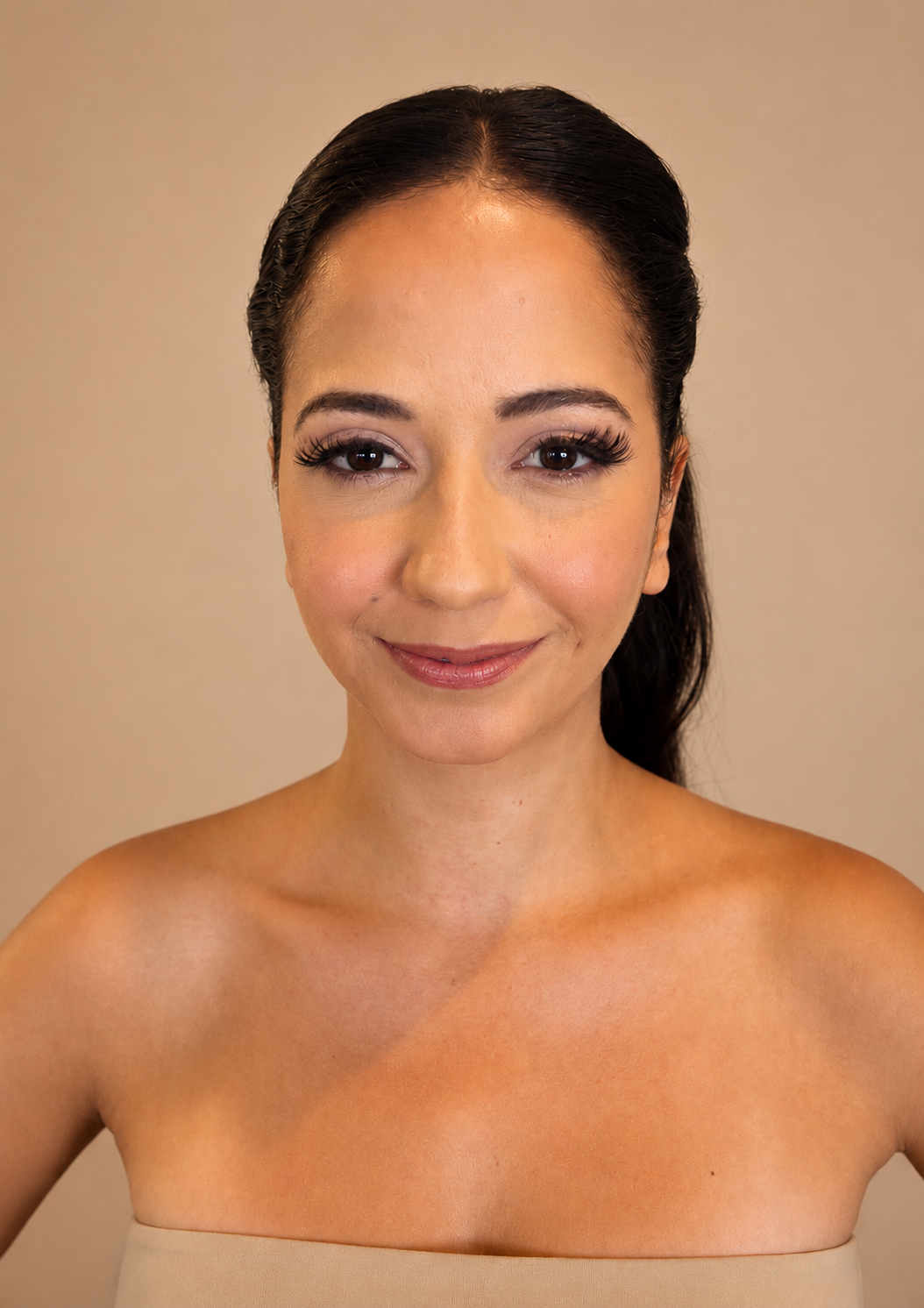 A woman with dark hair styled in a ponytail, wearing makeup, and smiling at the camera with a bare shoulder beige top against a plain background.