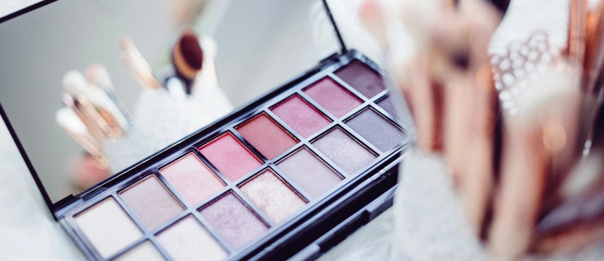 Close-up of a makeup eyeshadow palette with pink, purple, and neutral shades, placed near a mirror reflecting the palette and part of a person's face with earrings.