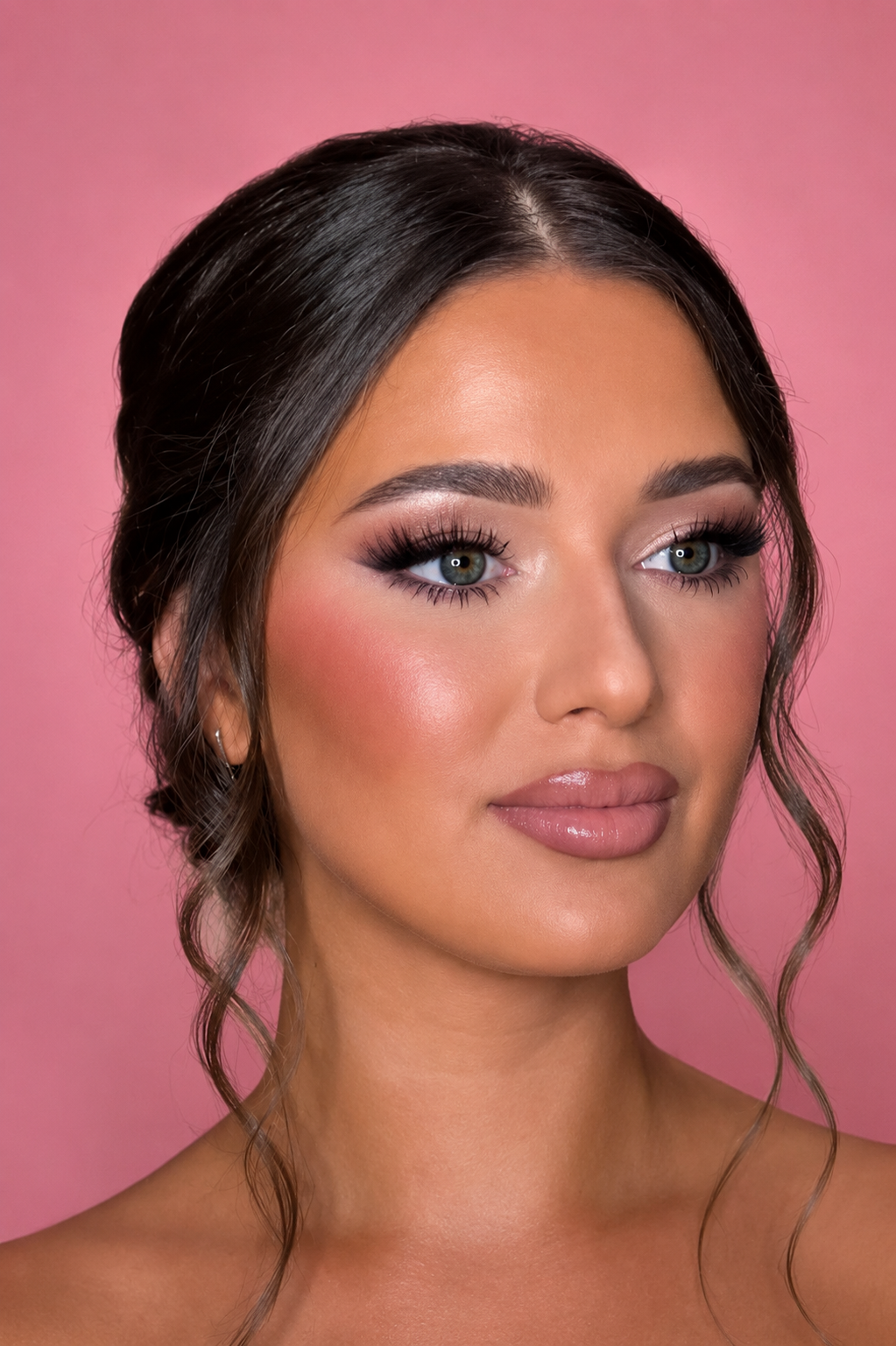 Close-up of a woman with dark brown hair styled in loose curls, makeup including false eyelashes, pink blush, nude lipstick, and a soft pink background.