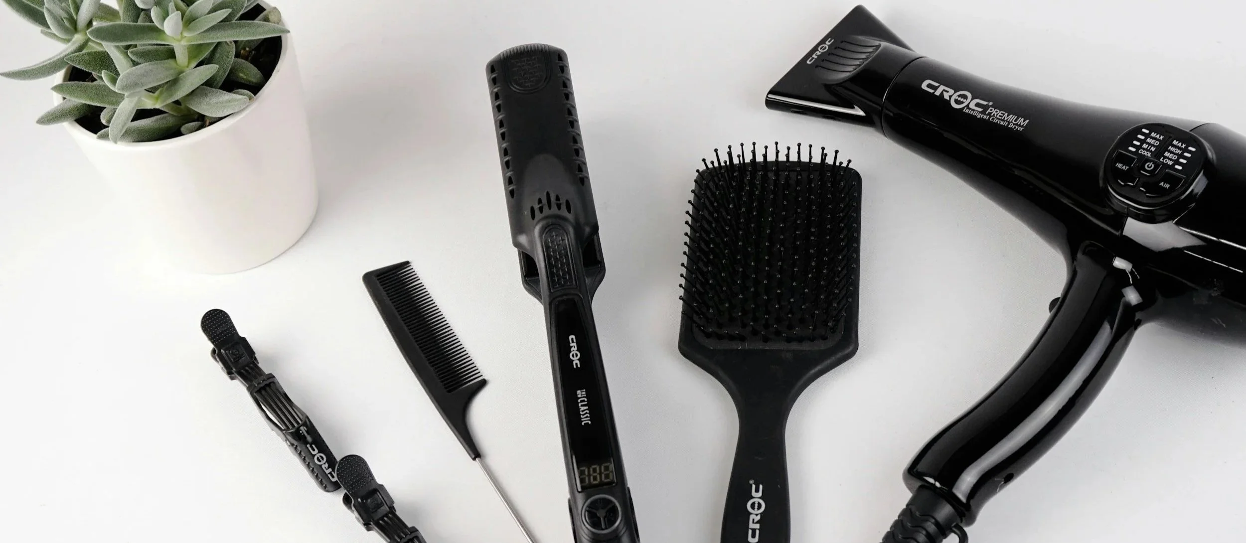 A collection of hair styling tools and a potted succulent on a white surface, including a hairdryer, hairbrush, comb, flat iron, curling iron, and hair clips.