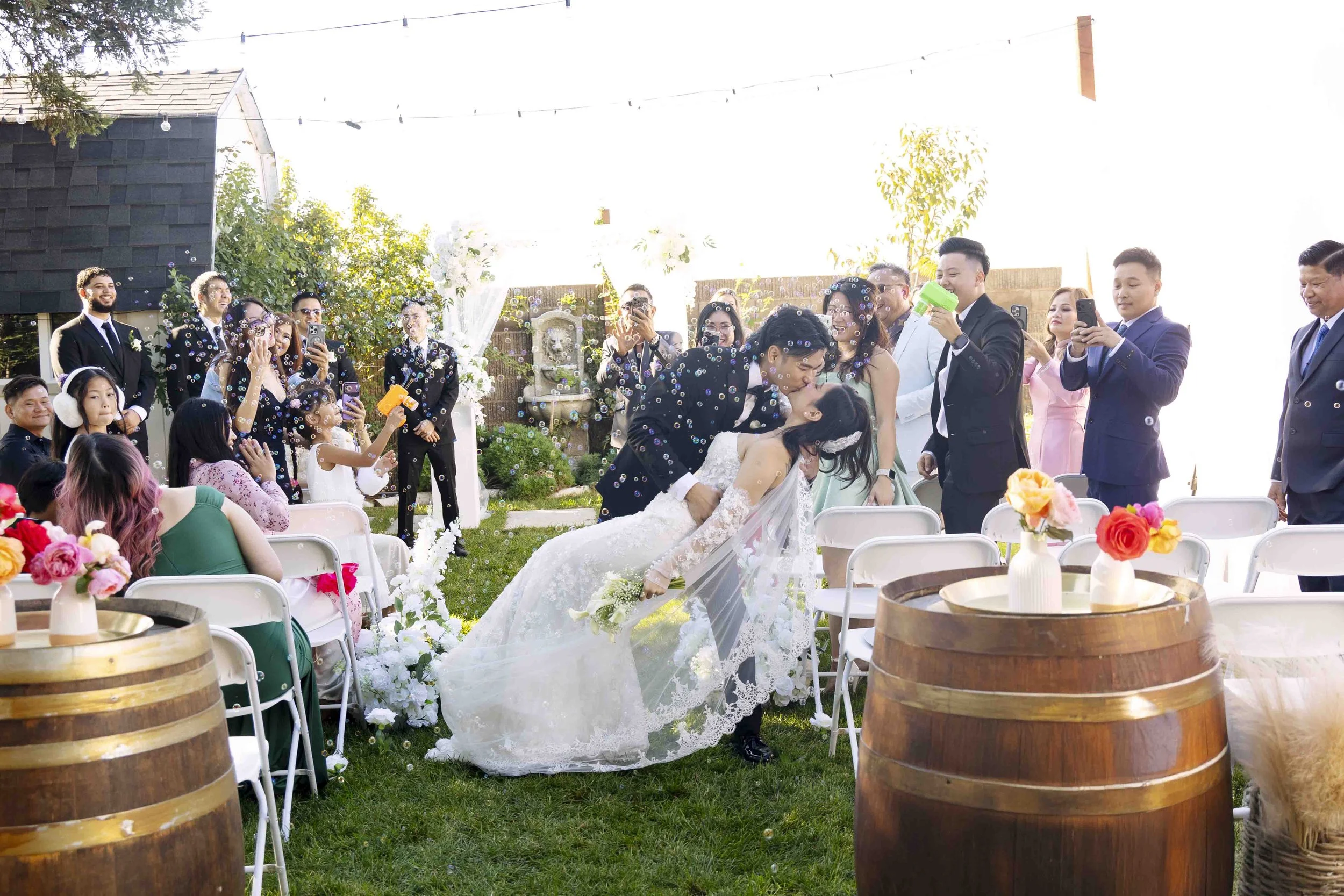 A wedding ceremony outdoors with the bride and groom kissing, surrounded by guests, some taking photos, with white chairs, floral arrangements, and a fountain in the background.