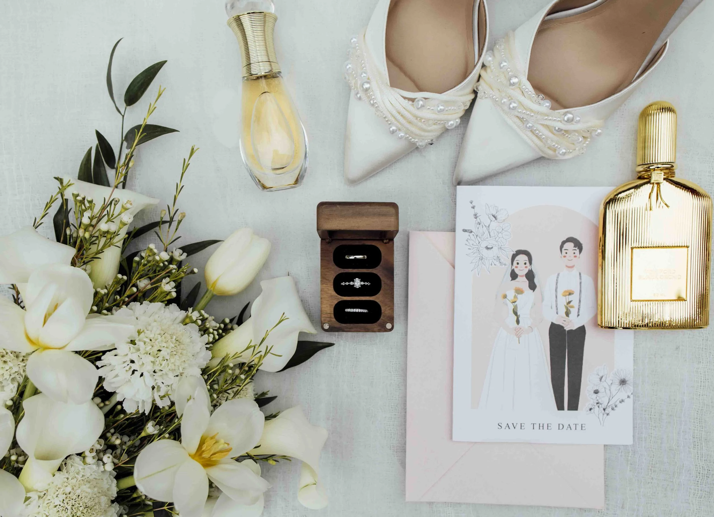 A bouquet of white flowers, a wedding invitation with illustrations of a bride and groom, perfume bottles, jewelry, and a sprig of greenery on a light-colored surface.
