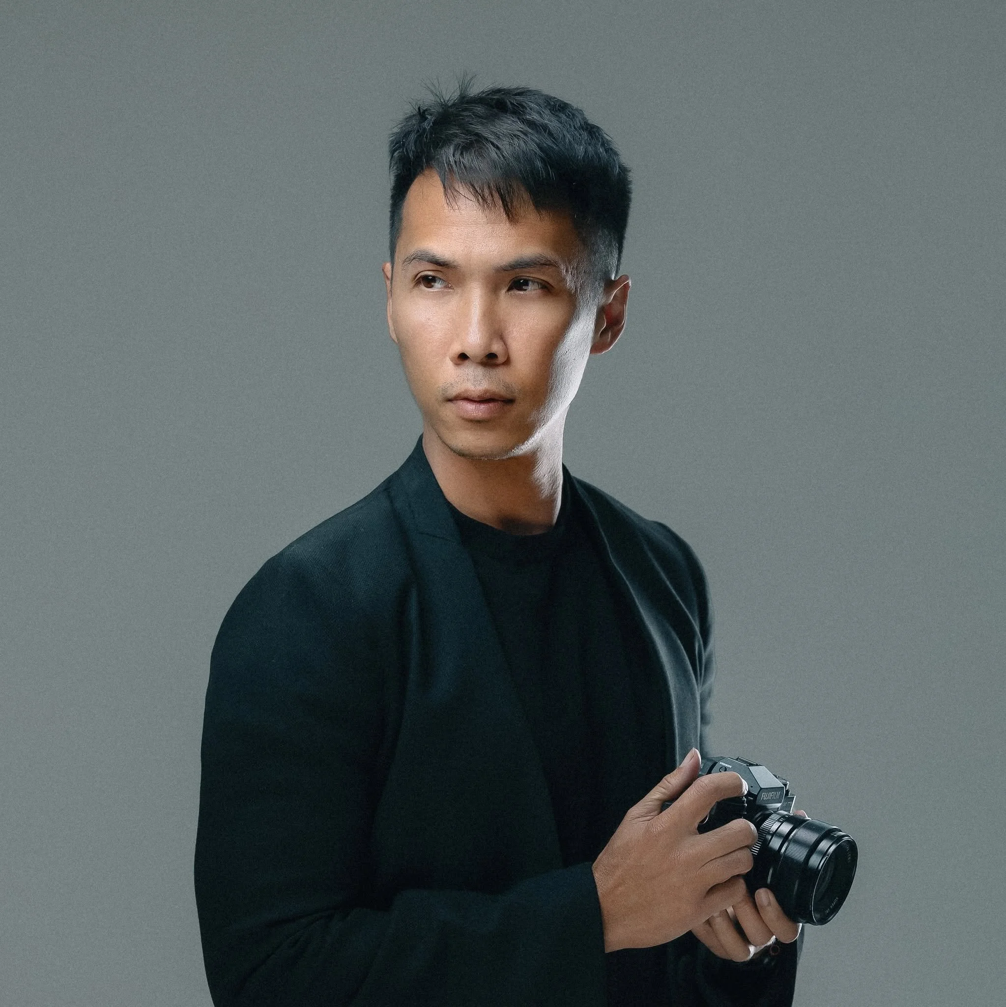 A young man with dark hair wearing a black suit, holding a camera and looking slightly to the side against a plain grey background.