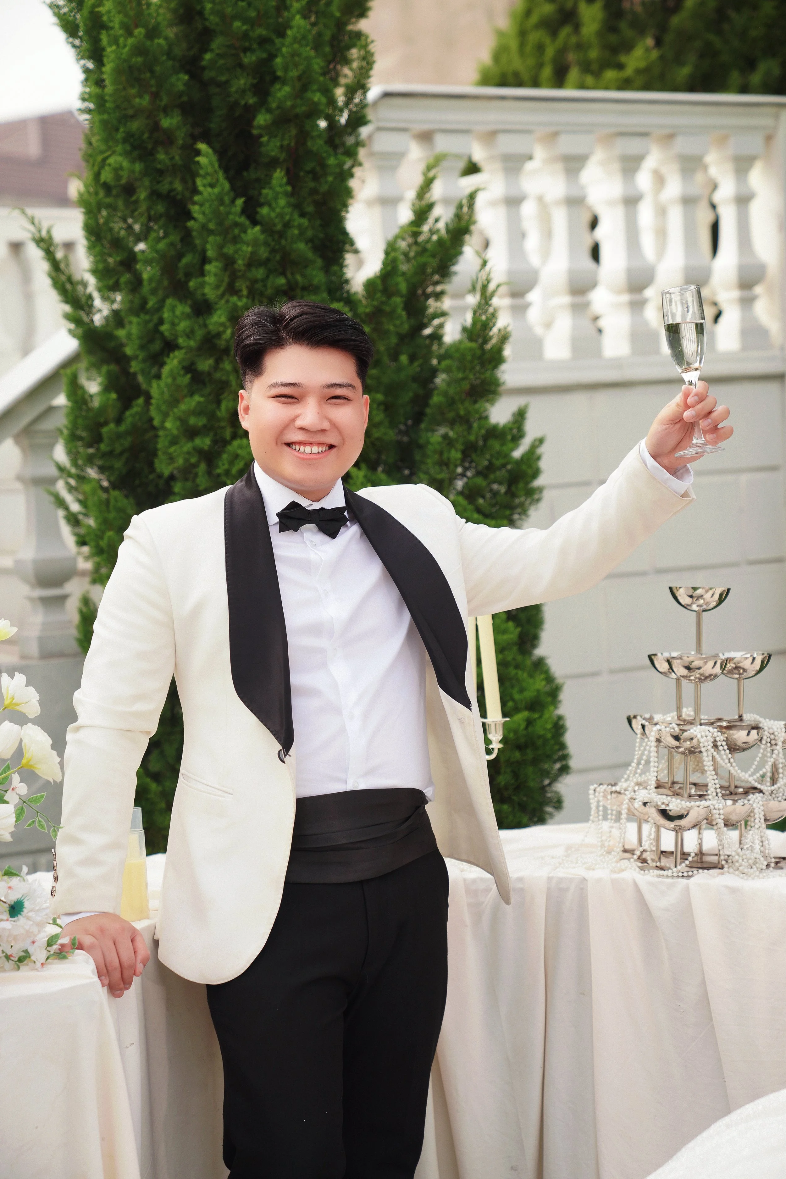 A man dressed in a white tuxedo jacket with black lapels, white shirt, and black pants, smiling and holding a champagne flute in an outdoor setting with greenery and a decorative table with a champagne tower.