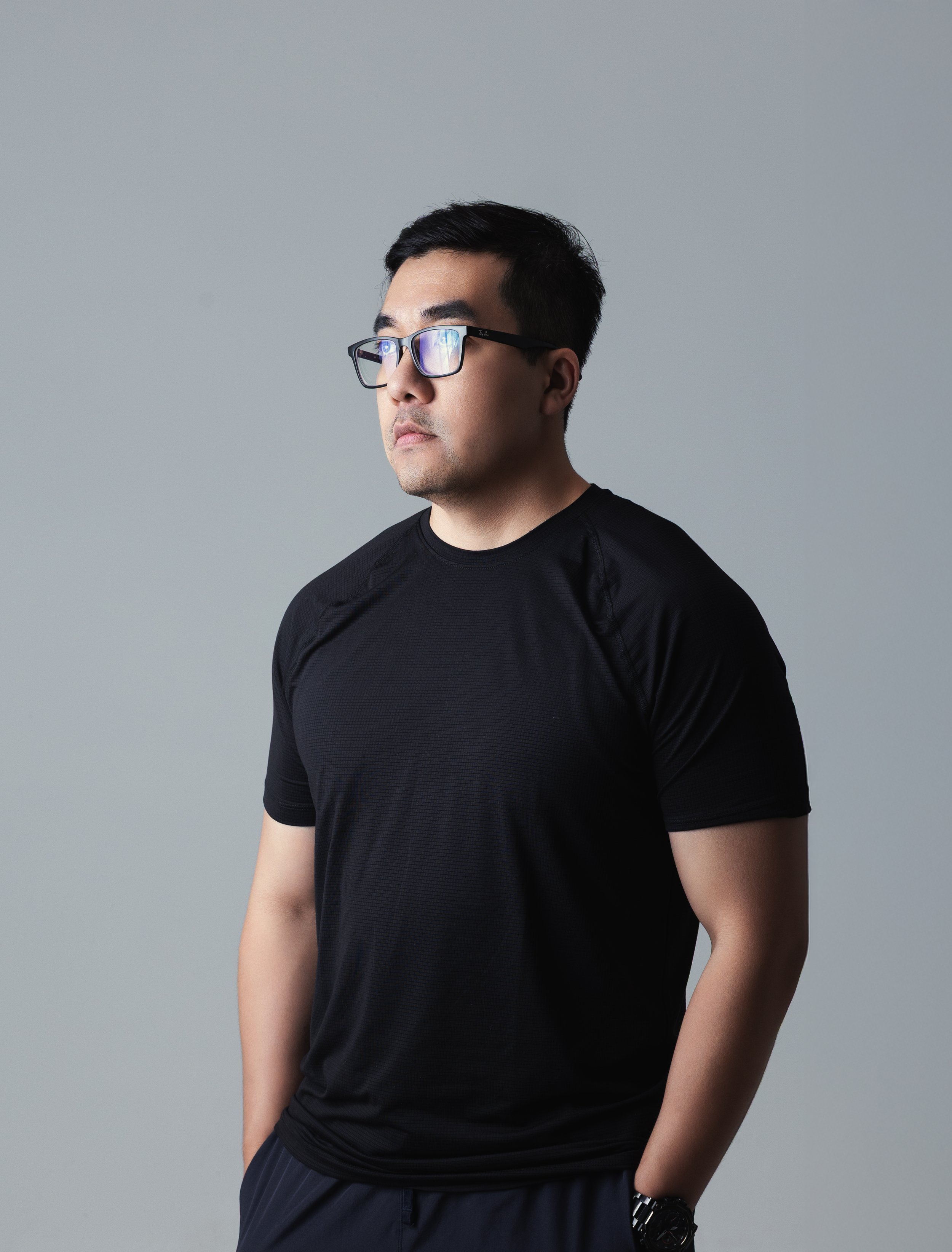 A young man with black hair and glasses wearing a black athletic shirt and watch, posing against a gray background.