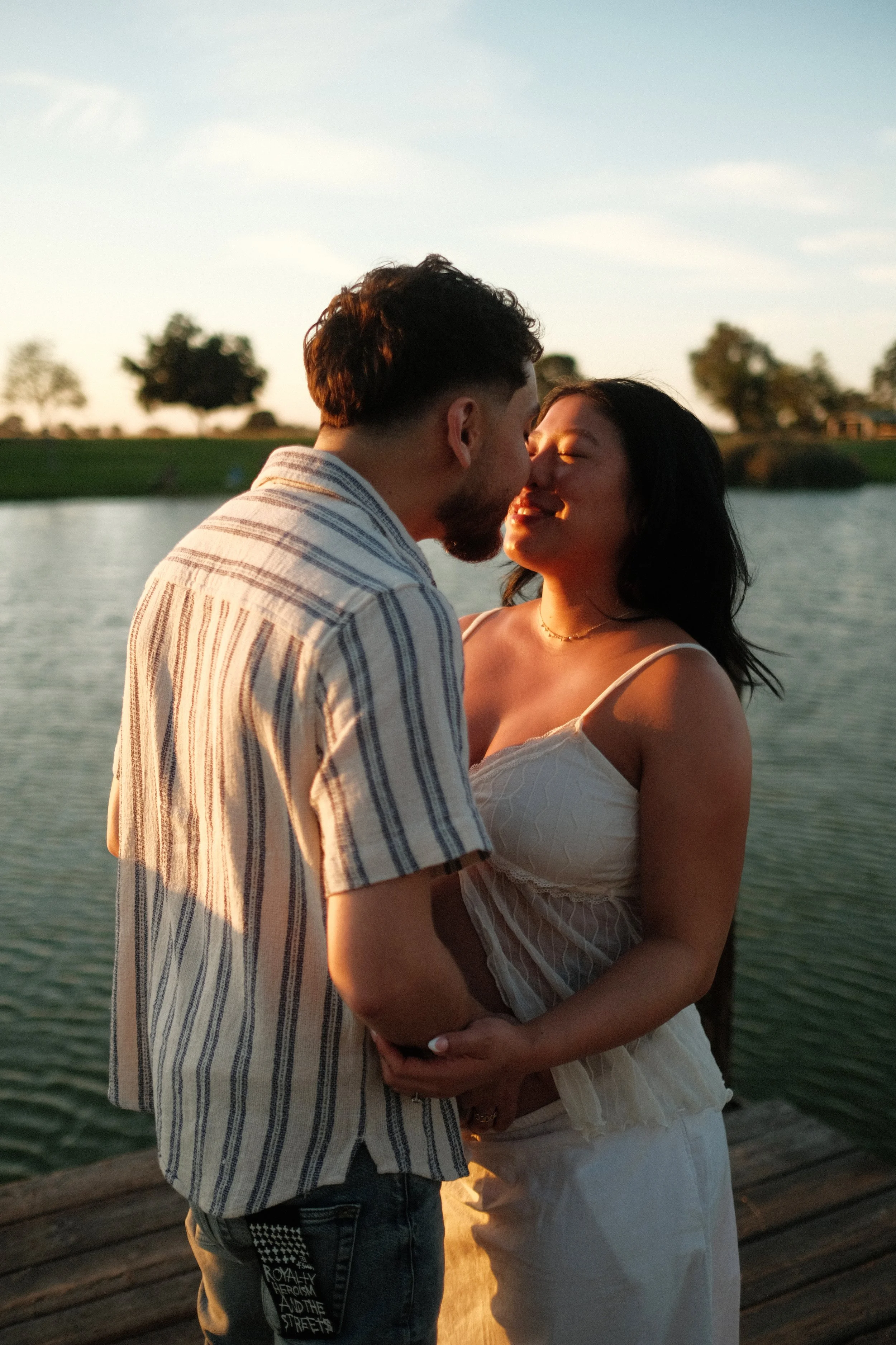 A young couple, a man and woman, stand close together near a body of water at sunset, about to kiss.