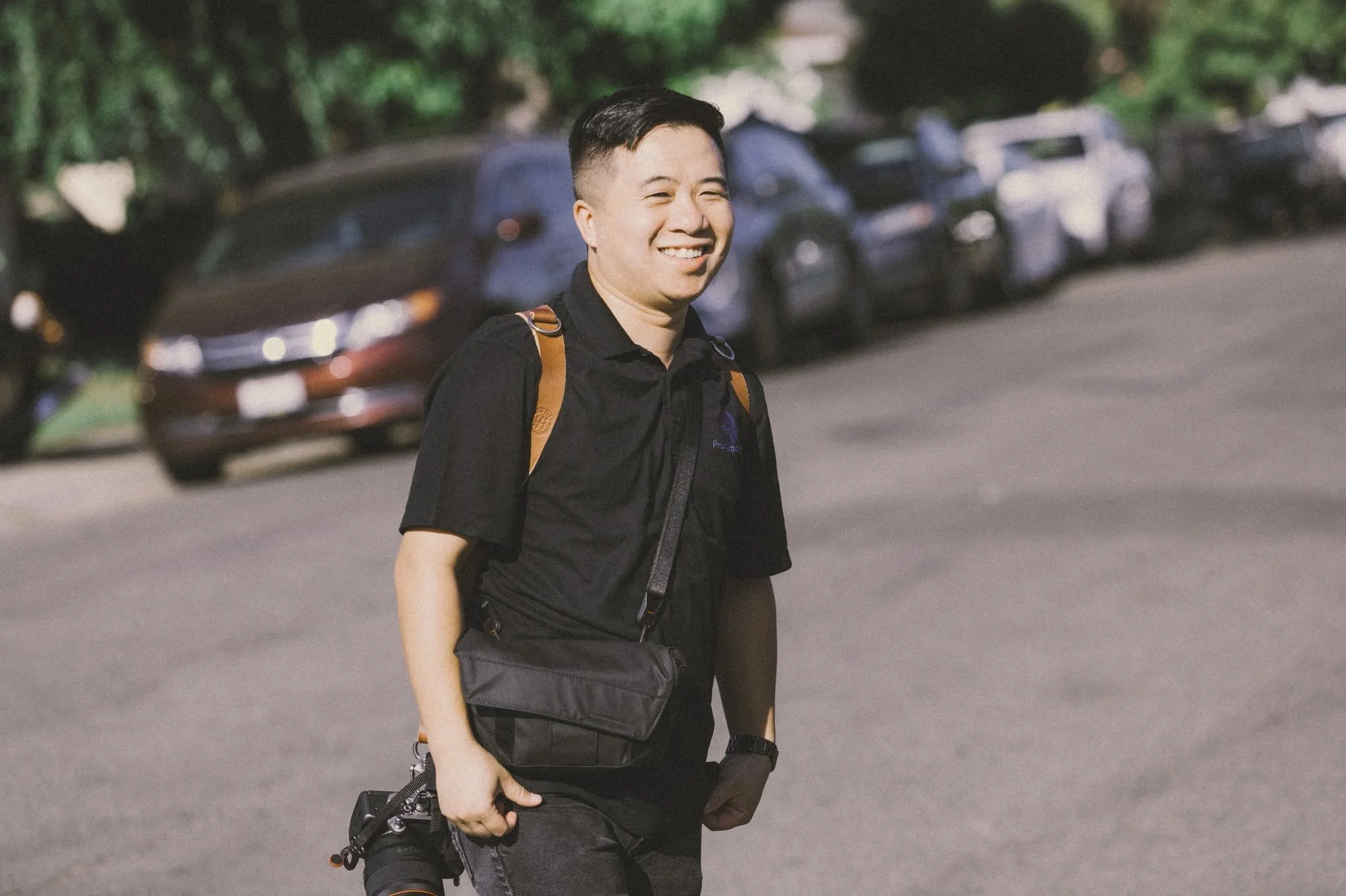 A young man with short dark hair smiling and walking outdoors with a background of parked cars and trees.