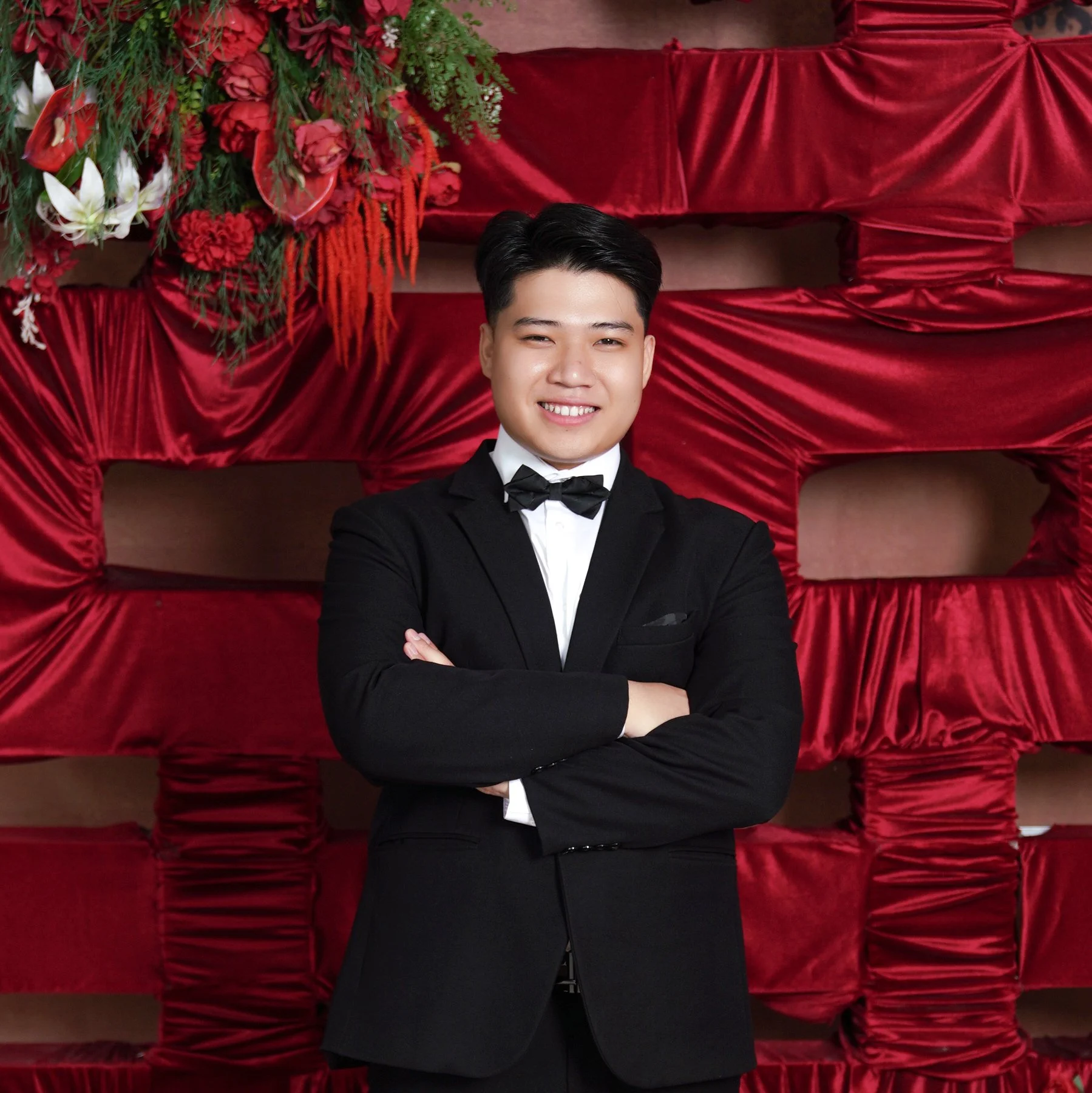 Young man in a tuxedo smiling with arms crossed, standing in front of a backdrop made of red velvet fabric with floral arrangements at the top left corner.