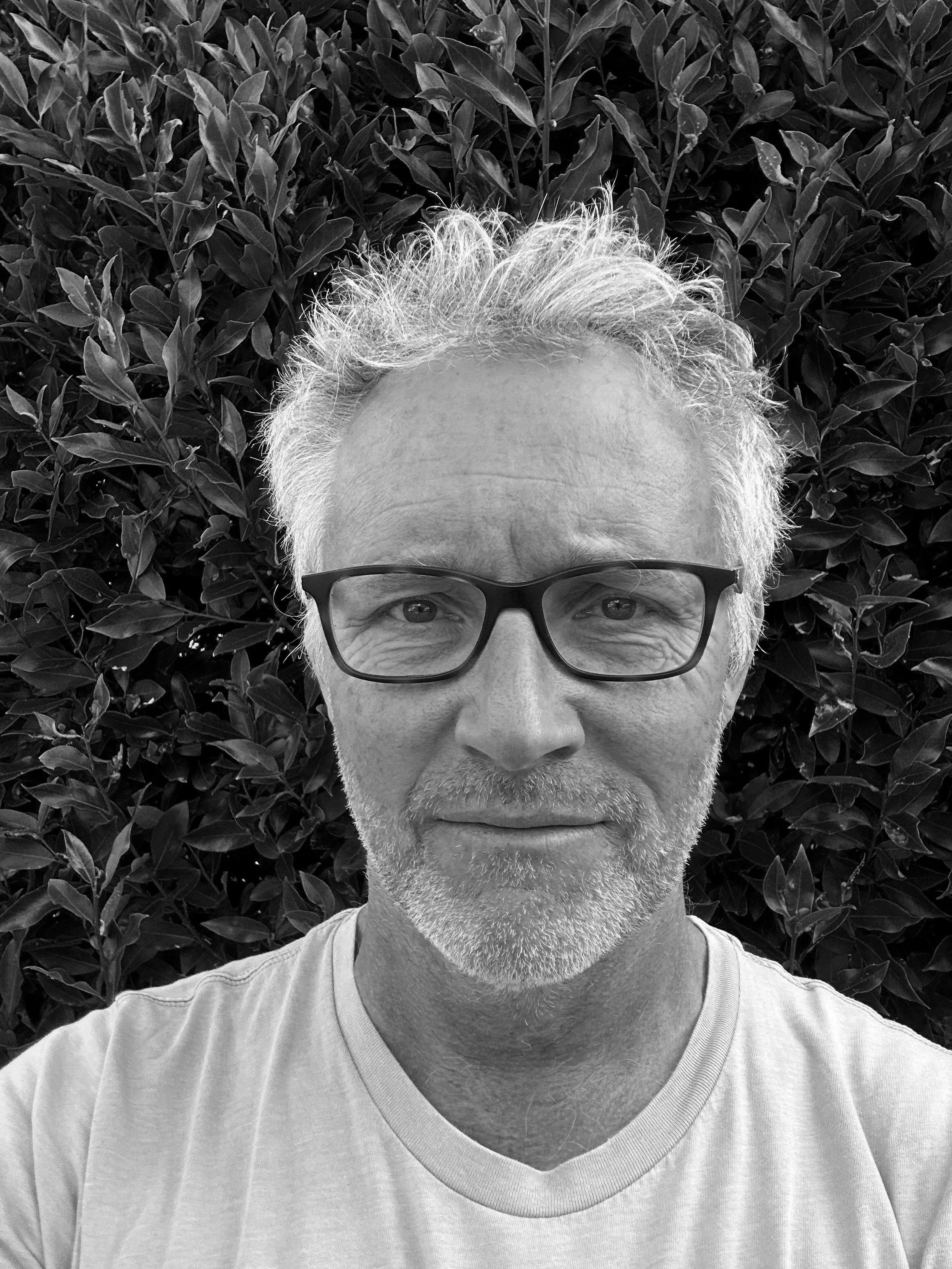 A black and white photo of a middle-aged man with light-colored hair, glasses, and a short beard, standing outdoors in front of dense leafy bushes, wearing a light-colored t-shirt.