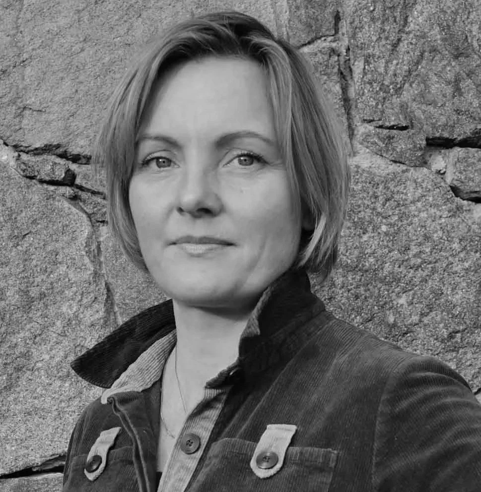 A woman with shoulder-length hair, wearing a dark jacket, standing outdoors in front of a stone wall.