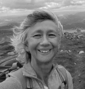 Smiling woman outdoors on a hillside with a scenic landscape, wearing a jacket and carrying a backpack.