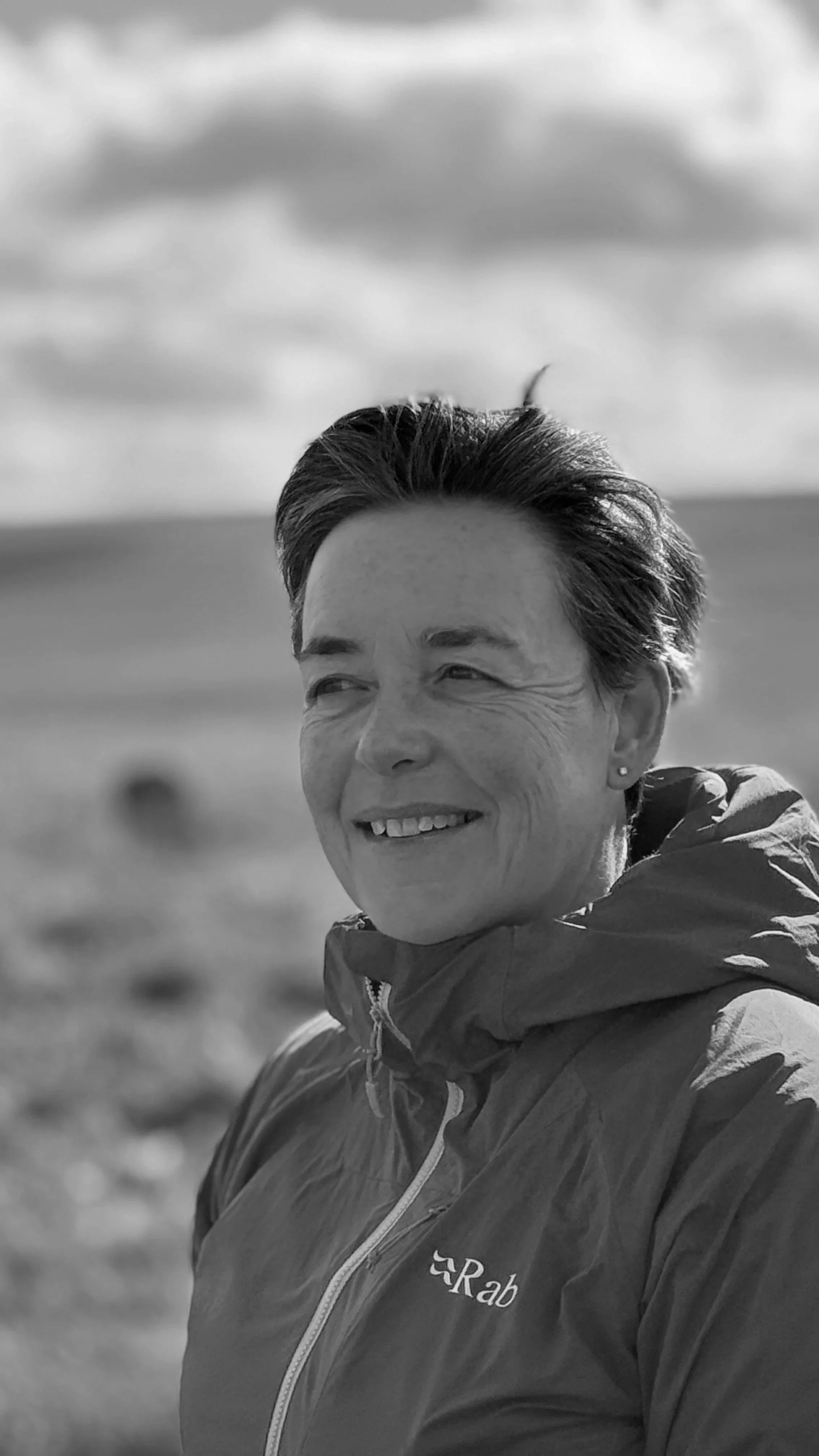 Black and white photo of a smiling woman outdoors wearing a jacket with the logo "Rab" on it, with a blurry landscape and cloudy sky in the background.