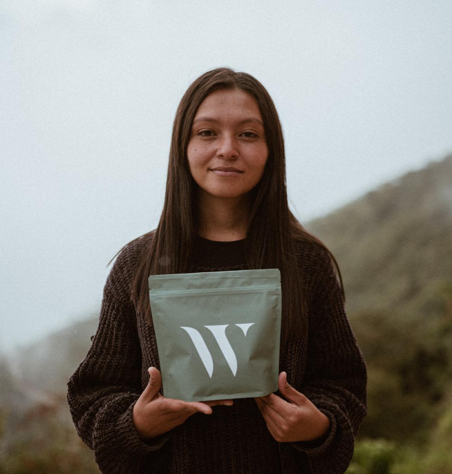 A woman standing outdoors holding KWC know where coffee W-label coffee beans package. She is standing in front of Colombia's mountainous landscape