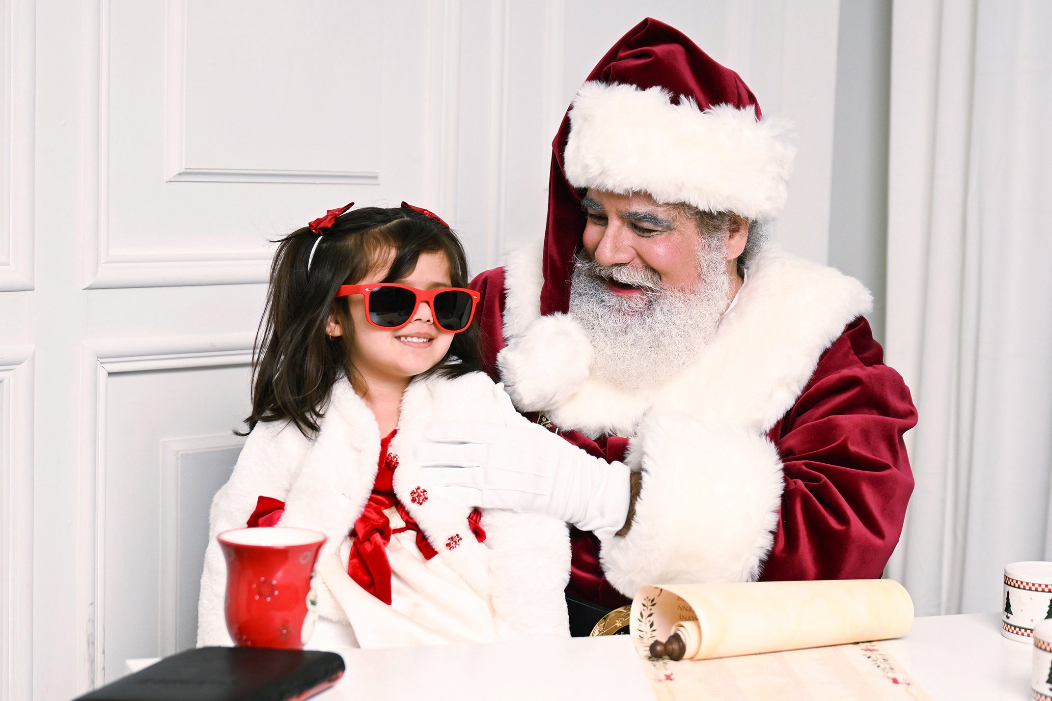 A girl wears a pair of red sunglasses while sitting on Santa Claus' lap.