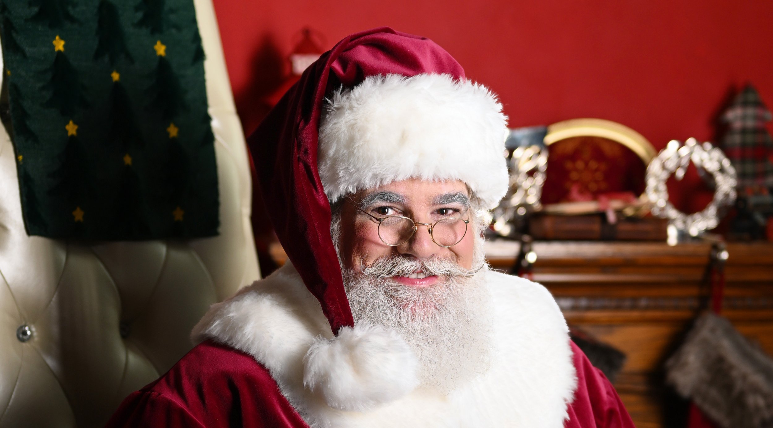 Santa Claus sitting on a cream-colored, tufted chair, wearing a red Santa hat and glasses, with a broad smile, in a decorated room with red walls and Christmas decorations.