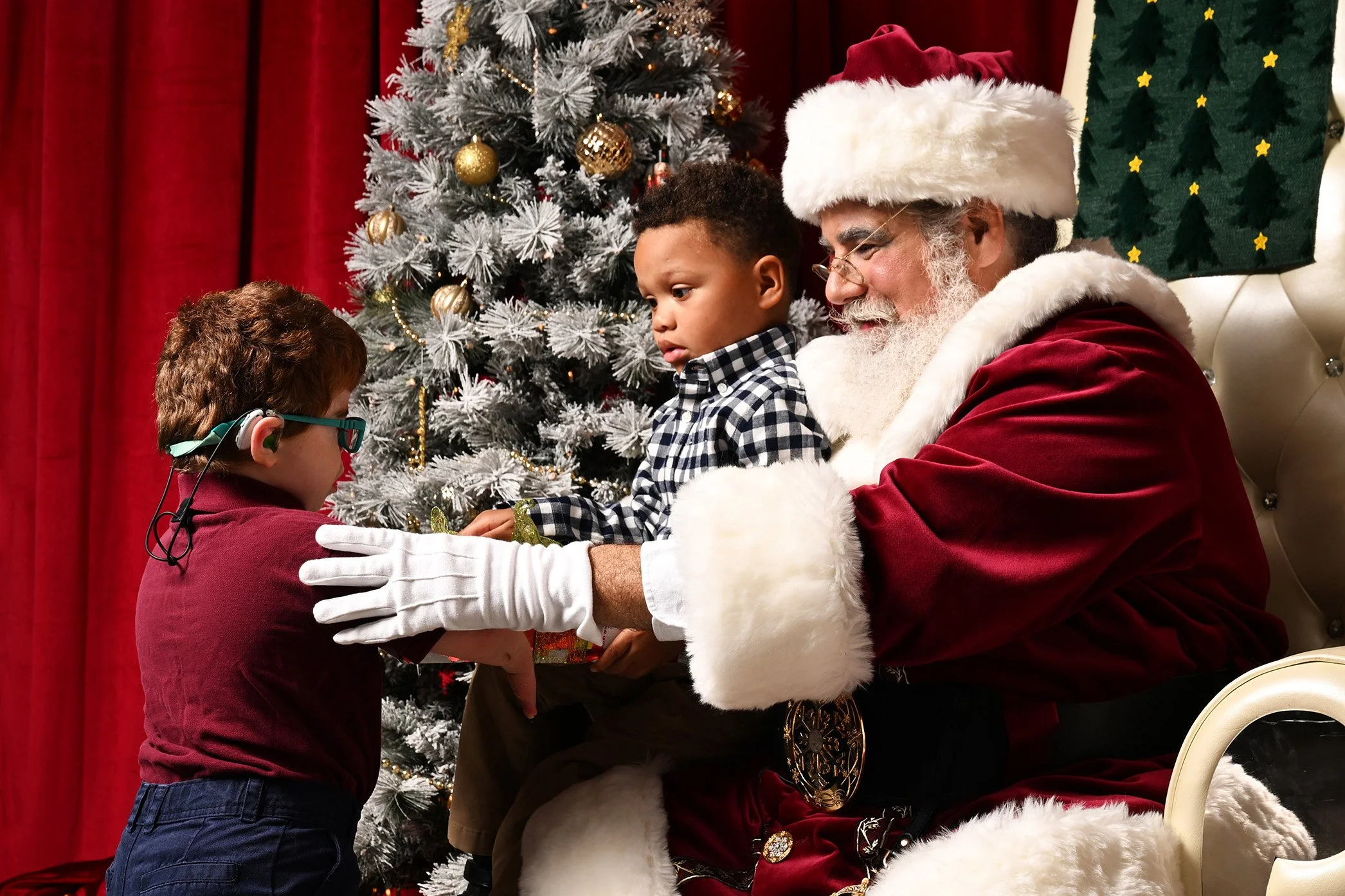 A young boy sits on Santa's lap as another boy approaches.
