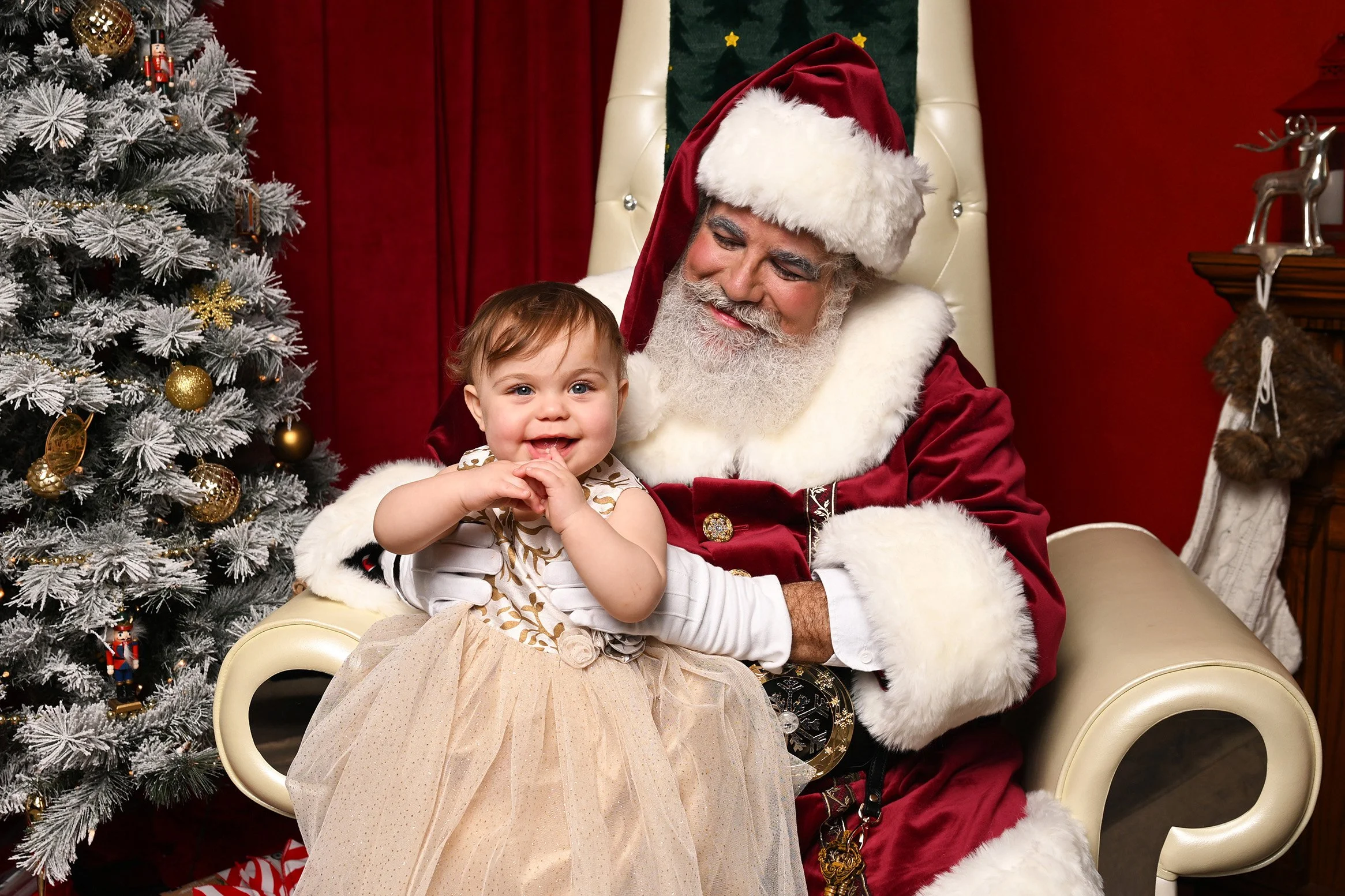 A toddler girl smiles and sits on Santa Claus' lap.