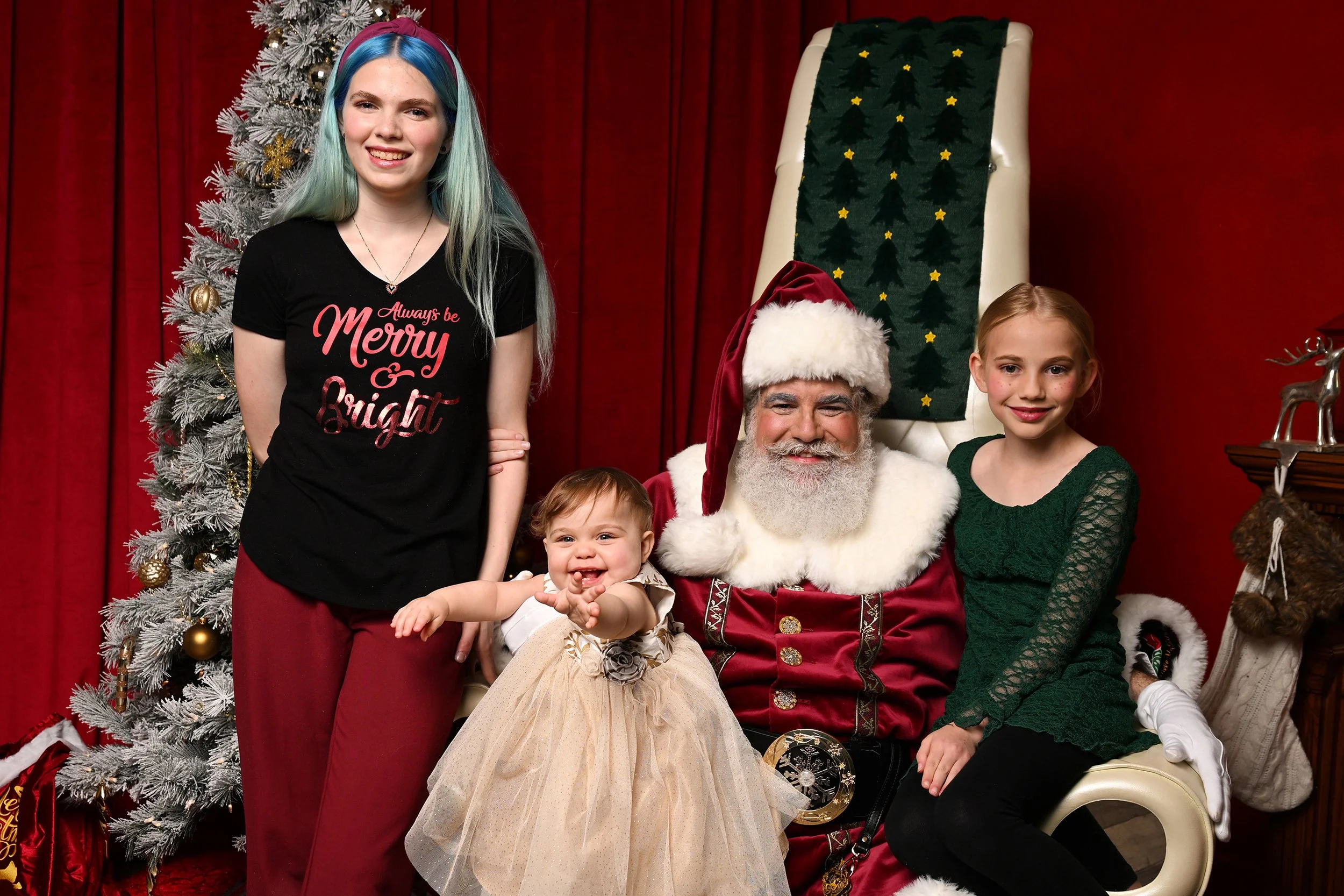 A group of children and Santa Claus posing in front of a Christmas tree and red curtains. The children are smiling and wearing festive clothing, with one girl in a green dress and another in a black shirt that says 'Always be Merry & Bright'.