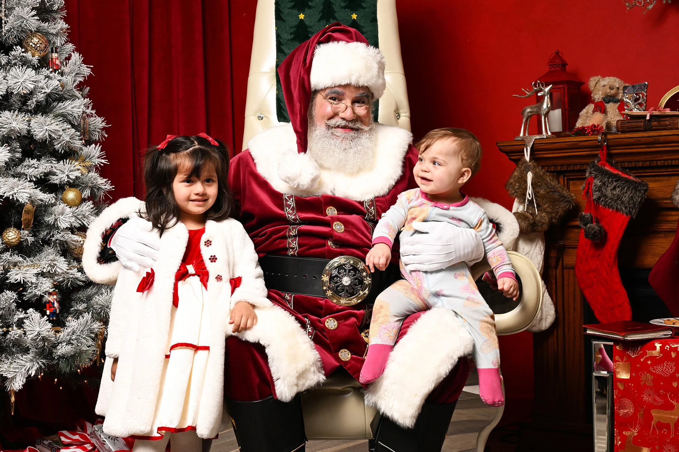 Santa Claus sitting in a white chair with two young girls, one on each side, during Christmas. A decorated Christmas tree is on the left, and a fireplace with stockings and holiday decorations is on the right, in a festive room with red walls.