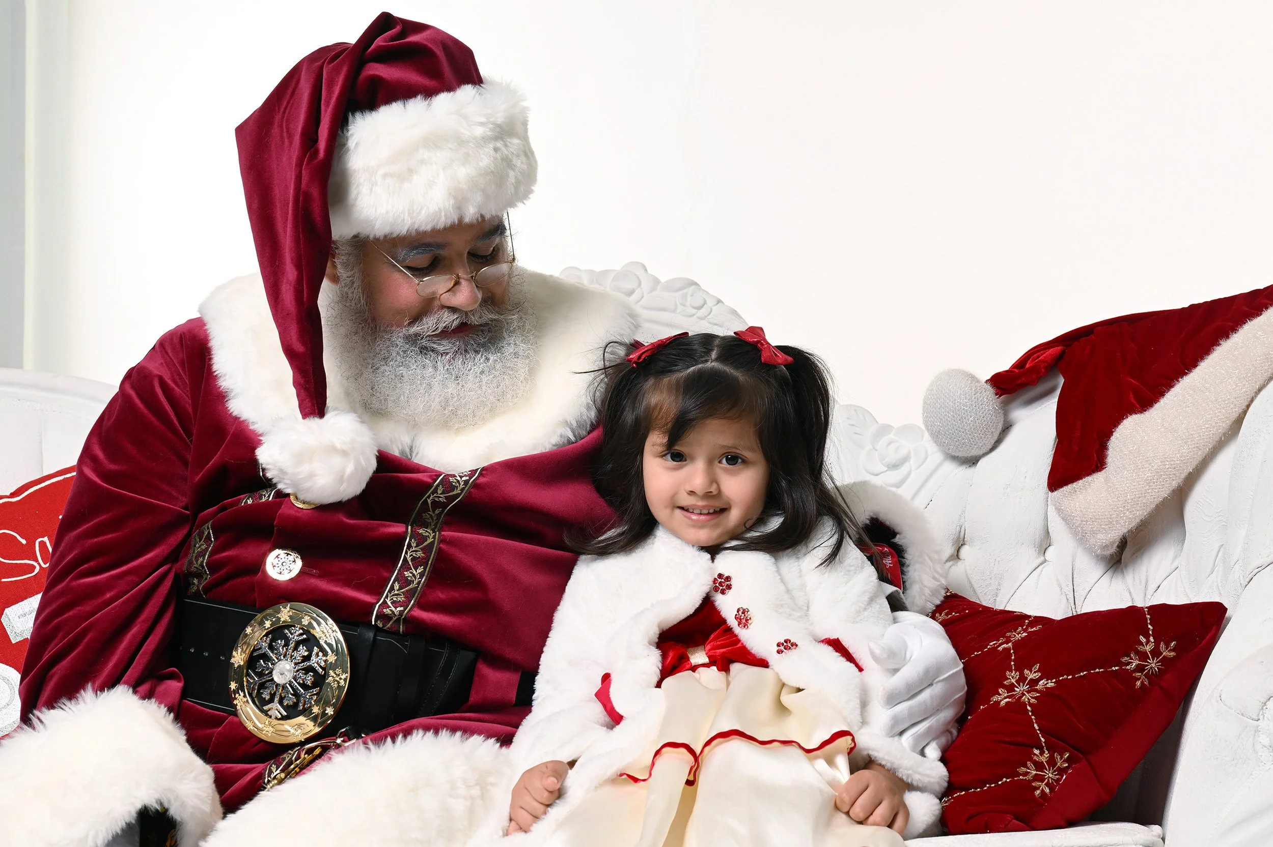 A smiling girl sits with Santa Claus on a white couch.
