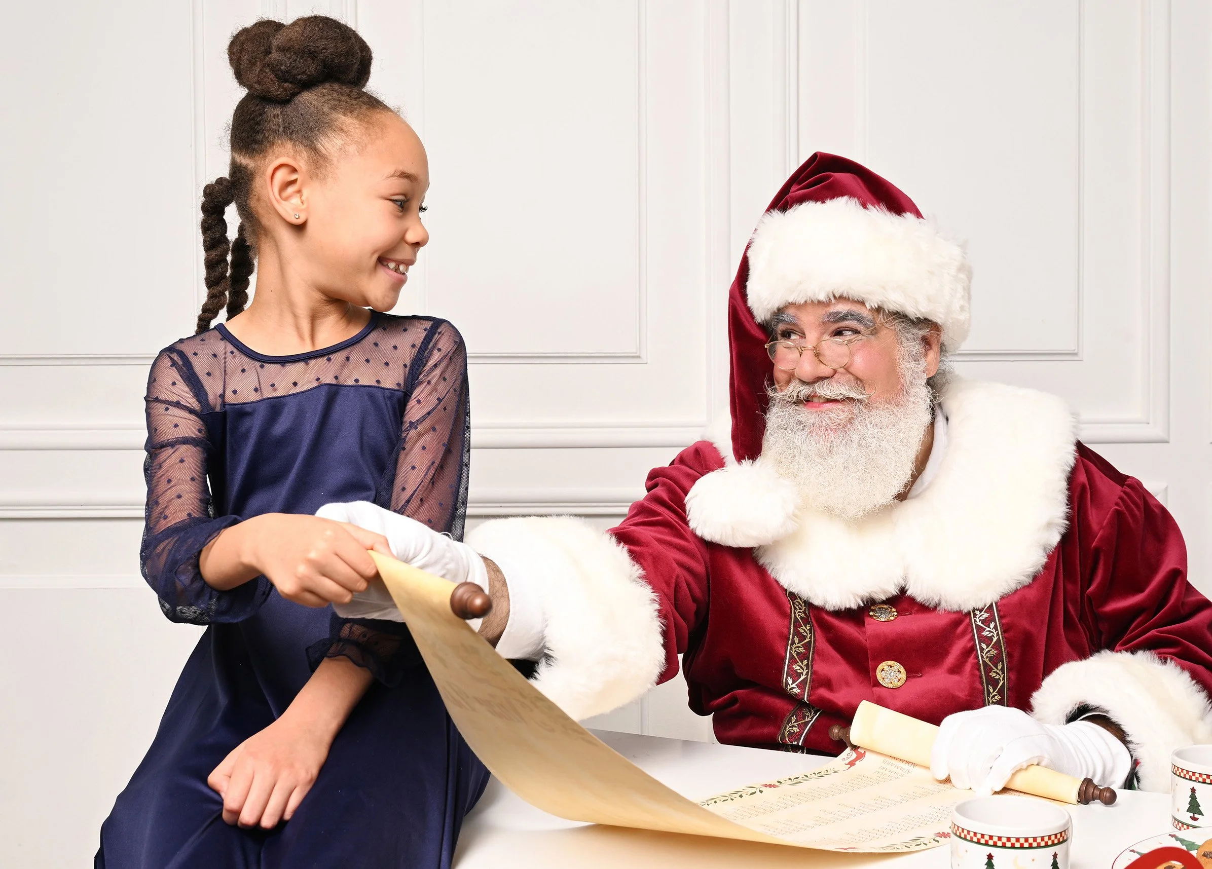 A young girl with a festive dress receives a scroll from Santa Claus, who is dressed in his traditional red and white outfit and hat. They are indoors against a white paneled wall, and there are Christmas-themed cups and decorations on the table.