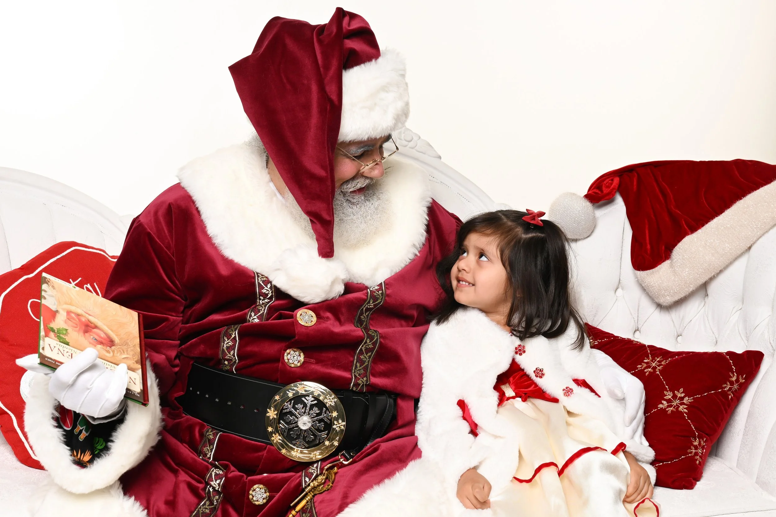 Santa Claus dressed in red velvet suit with white fur trim, sitting on a white sofa with a young girl, both looking at each other. Santa is holding a small book and has a round decorated belt buckle and a red stocking with white fur trim next to him. The girl wearing a white and red Christmas outfit has a red bow in her hair and is smiling.
