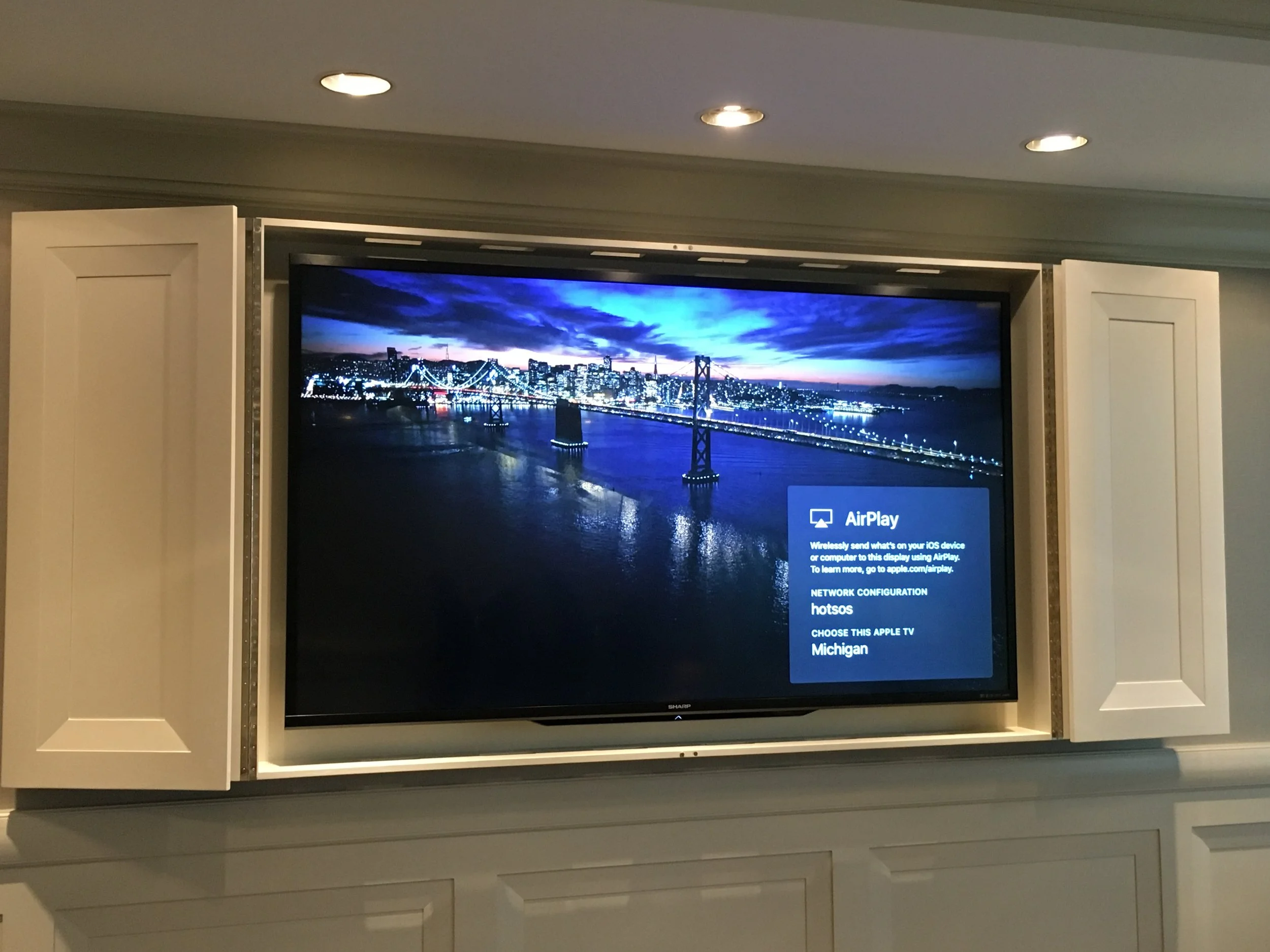 A wall-mounted flat-screen television displaying a cityscape at night, with bridge lights and a dark cloud-filled sky, partially obscured by open white cabinet doors on either side.