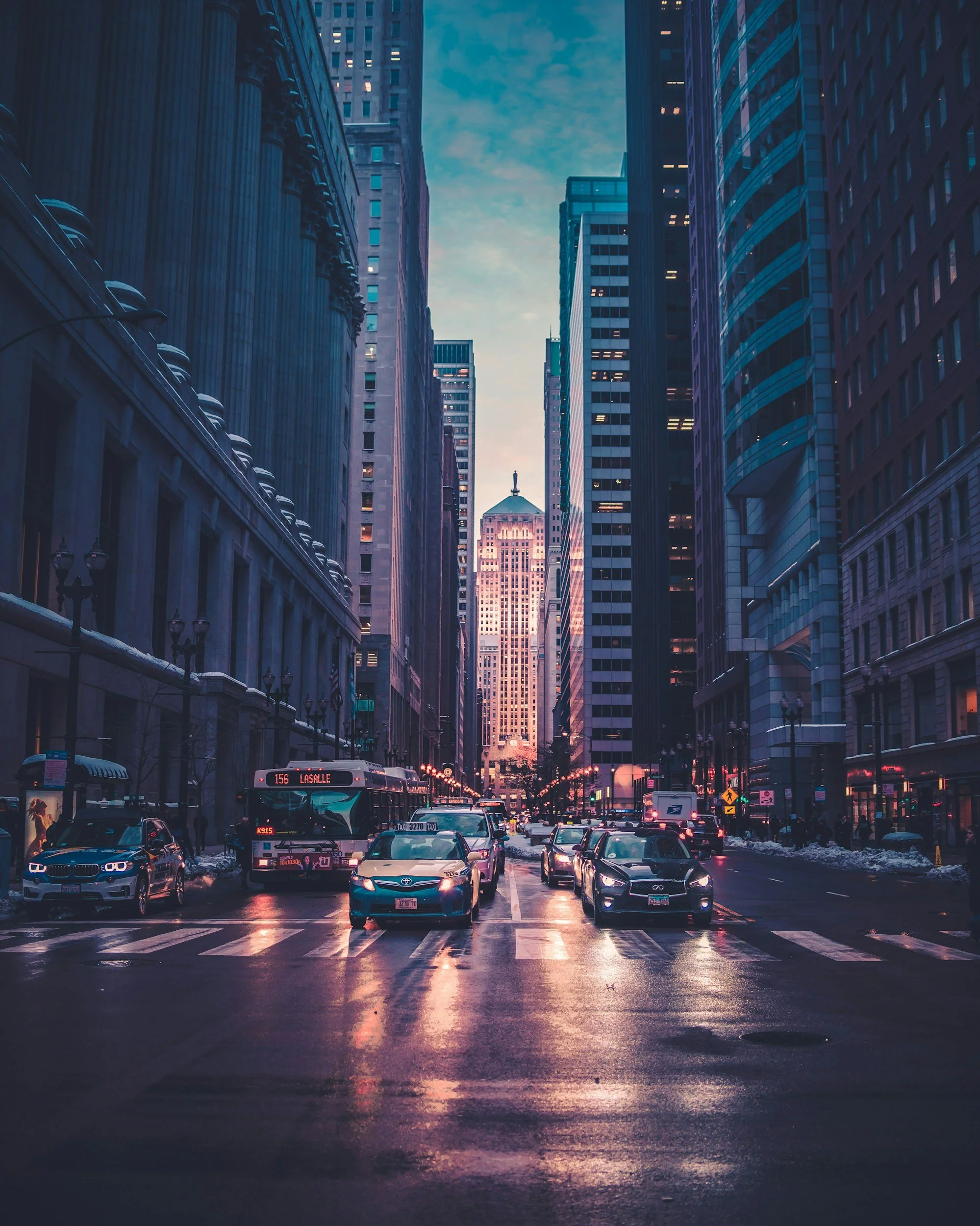City street scene at dusk with tall skyscrapers on both sides, cars, buses, and taxis on the wet road, with a view of a historic building in the distance and a cloudy sky overhead.