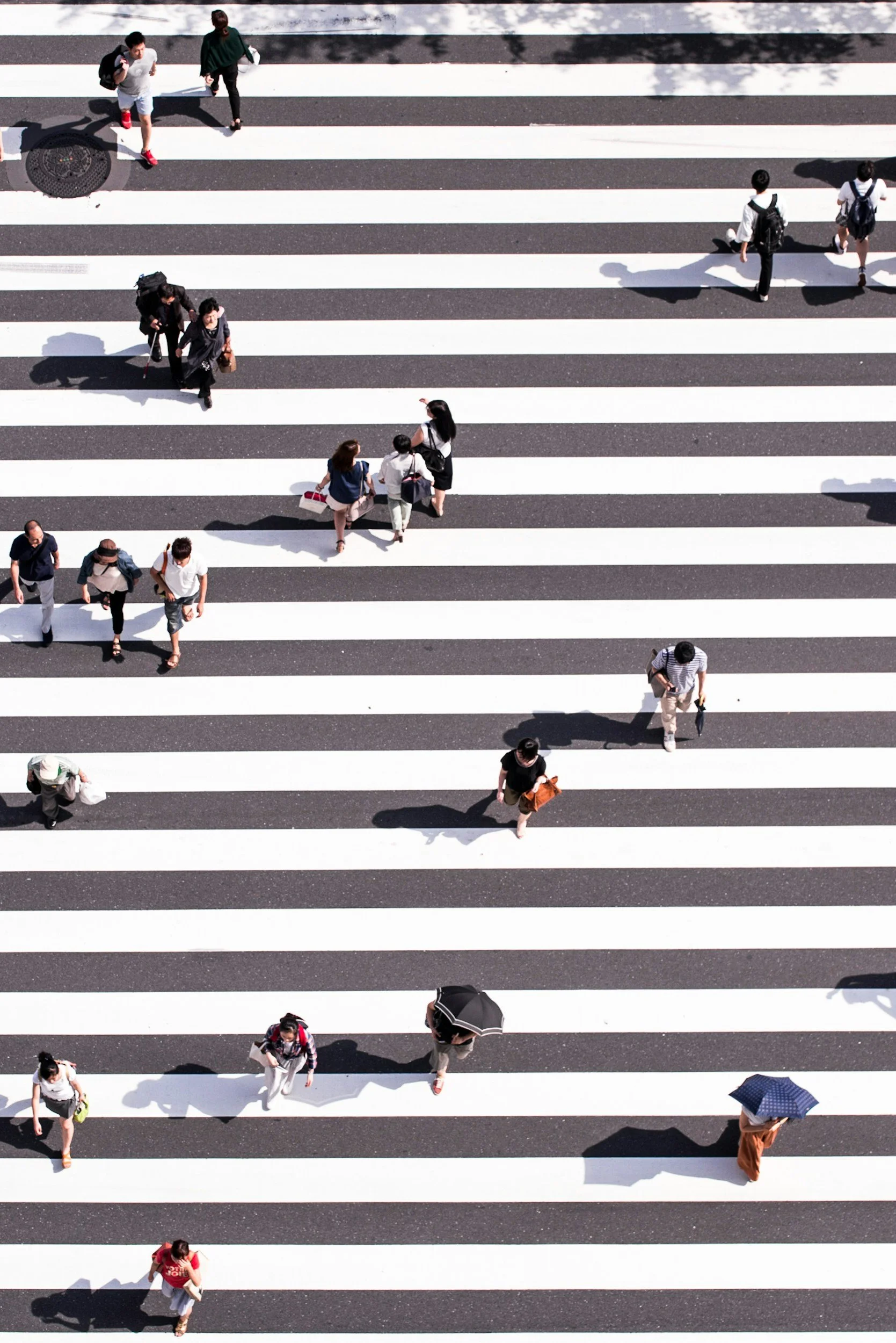 People walking across a pedestrian crosswalk on a sunny day, some carrying umbrellas and backpacks.
