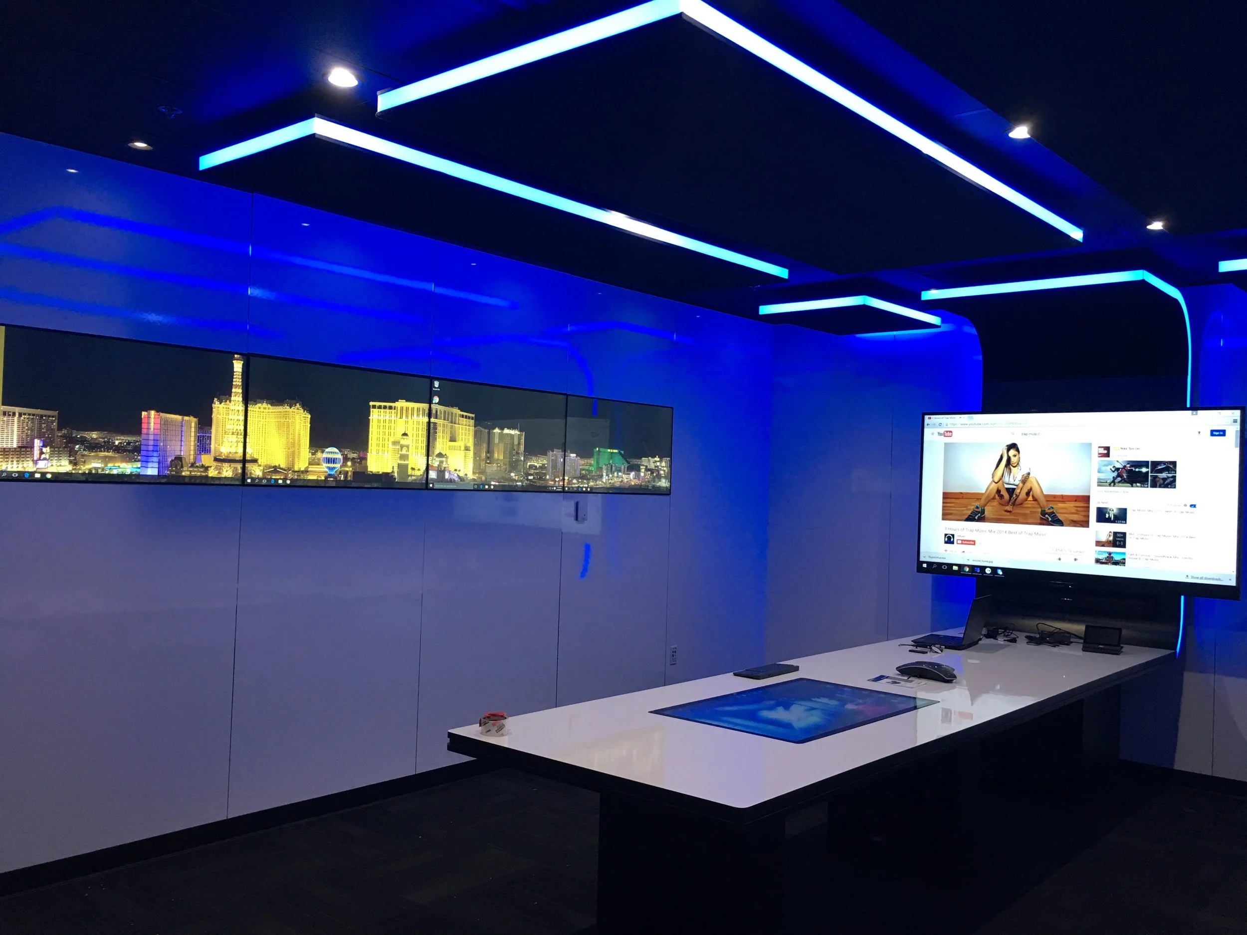 An indoor space with blue lighting fixtures on the ceiling, a large window showing a cityscape at night, and a computer monitor displaying a YouTube video of a woman sitting on the floor.