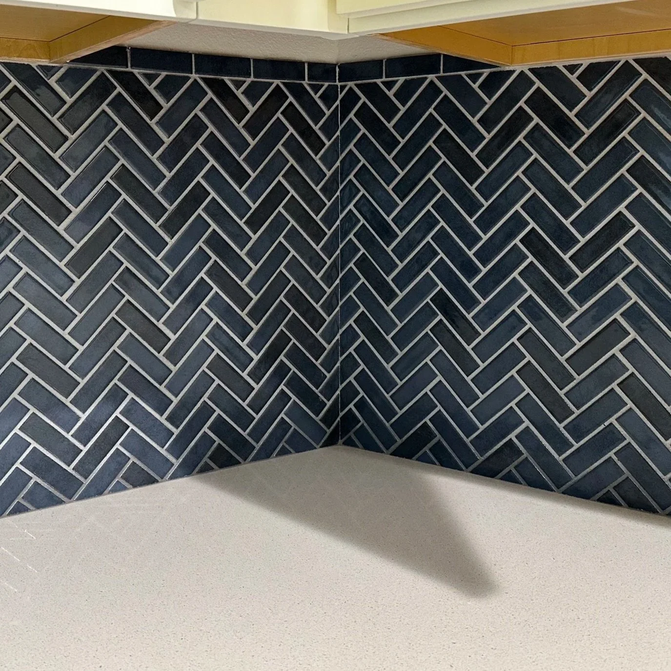 Close-up of a kitchen counter with a black herringbone tile backsplash, a white electrical outlet, and part of a white stove and upper cabinets.